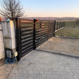 LDR Electrical Dariusz Karaś - Nowoczesna, czarna brama wjazdowa z automatyką, widoczna z perspektywy kostki brukowej. W tle drzewo bez liści i zalesione wzgórza o zmierzchu.