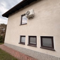 LDR Electrical Dariusz Karaś - Boczny widok domu z zamontowaną jednostką klimatyzacji na ścianie, nad oknem. Widoczne trzy okna na dole i chodnik z kostki brukowej.