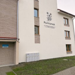 LDR Electrical - Elewacja budynku 'Rumianek' z widocznymi skrzynkami elektrycznymi na zewnątrz. Ujęcie z poziomu chodnika, zieleń wzdłuż budynku.