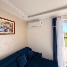 LDR Electrical - Biały klimatyzator ścienny w jasnym salonie z niebieską sofą narożną i obrazem kwiatów. Widok na zewnątrz przez okno z firankami.