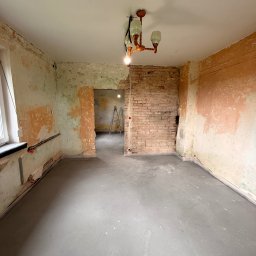 LDR Electrical - Surowe wnętrze w trakcie remontu: świeżo wylana posadzka, odsłonięta cegła, instalacja elektryczna w ścianie. Widoczna drabina w przejściu do kolejnego pomieszczenia.
