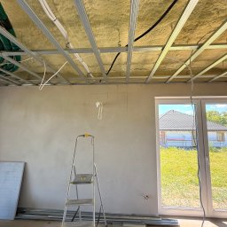 LDR Electrical - Montaż instalacji pod sufitem podwieszanym z izolacją, widoczne kable i rury. W tle drabina i okno z widokiem na dom w budowie.