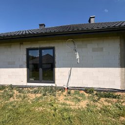LDR Electrical - Ściana budynku z pustaków z oknem i wyprowadzonymi kablami. Widoczne elementy instalacji elektrycznej na zewnątrz. Trawa i ziemia w dolnej części kadru.