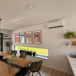 LDR Electrical - Biały klimatyzator ścienny w nowoczesnym wnętrzu z drewnianym stołem, krzesłami i oknem z widokiem na zieleń. Na ścianie wiszą obrazy w ramkach.