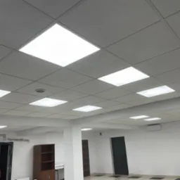 Sufit podwieszany z kwadratowymi panelami oświetleniowymi LED w biurze, widoczna instalacja elektryczna przy drzwiach i pracownik w pomarańczowej koszulce.