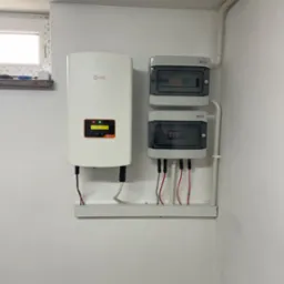 Biały inwerter Solis zamontowany na ścianie obok dwóch szarych rozdzielnic elektrycznych z widocznymi bezpiecznikami, instalacja w rogu pomieszczenia z białymi ścianami i rurami.