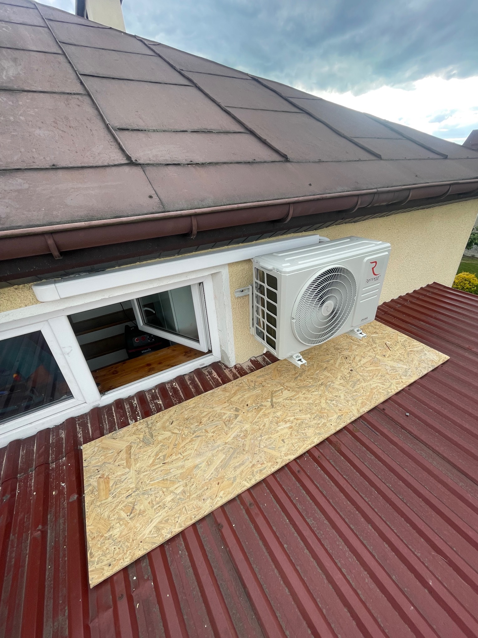 Biały klimatyzator Rotenso zamontowany na elewacji domu, widoczny z dachu pokrytego blachą trapezową i płytą OSB. Widok z góry, pochmurne niebo.
