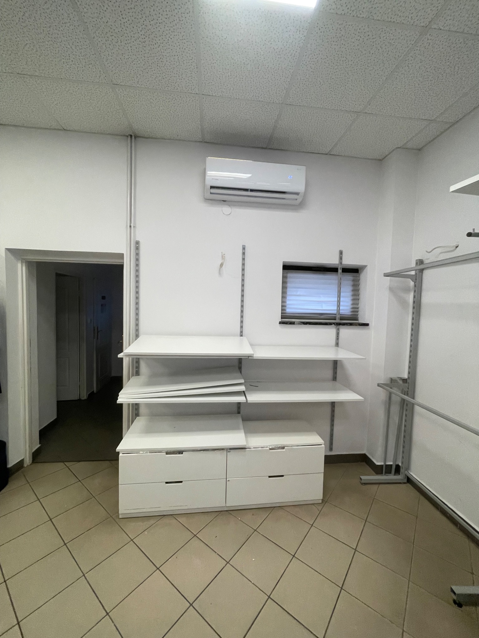 Biała klimatyzacja ścienna nad oknem z żaluzjami w jasnym pomieszczeniu z regałami i szafkami. Widok na korytarz w tle. Minimalistyczny wystrój.