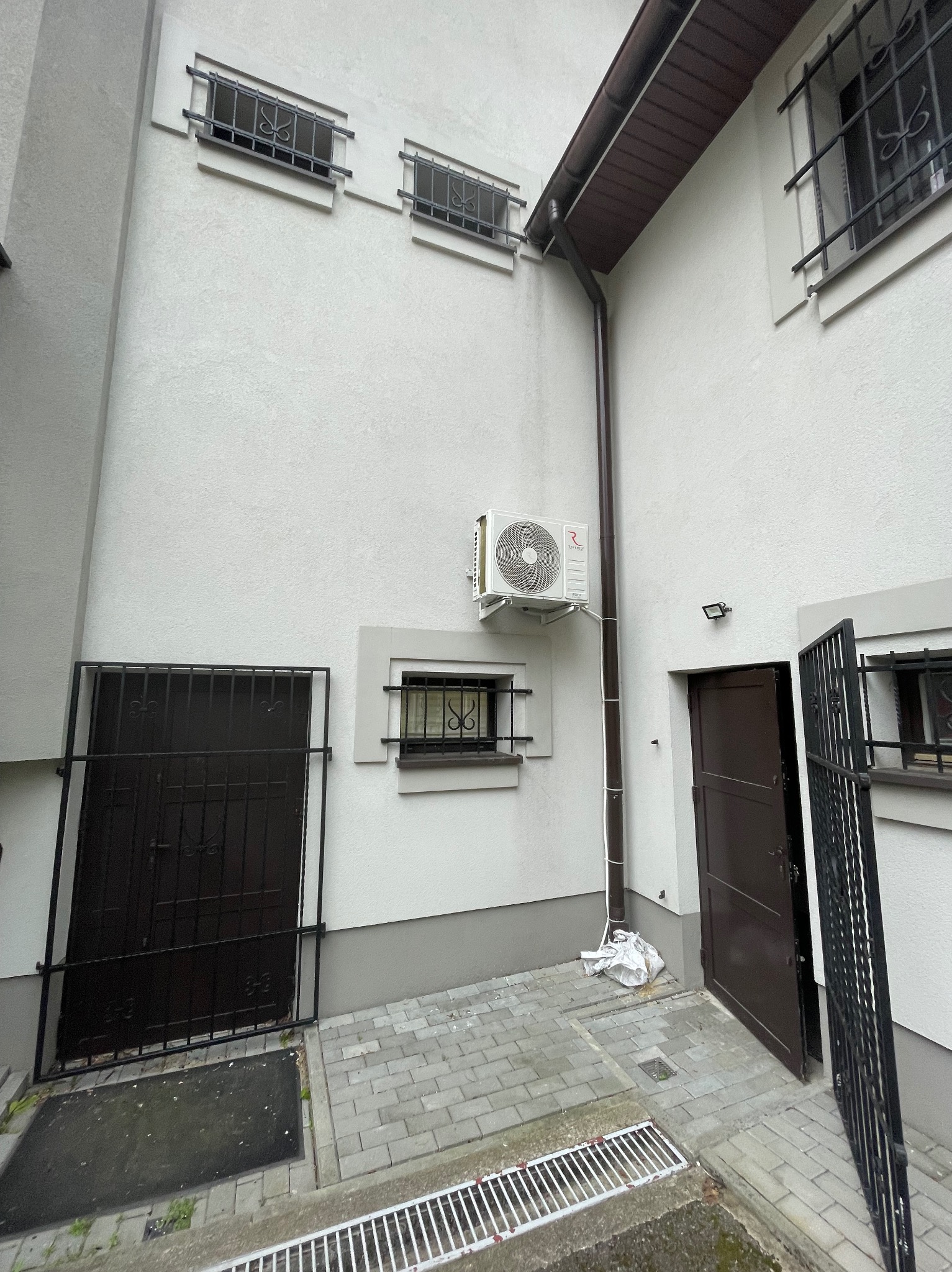 Biała elewacja budynku z zamontowaną jednostką zewnętrzną klimatyzacji, brązową rynną i kratami w oknach. Widoczne drzwi wejściowe i fragment chodnika.
