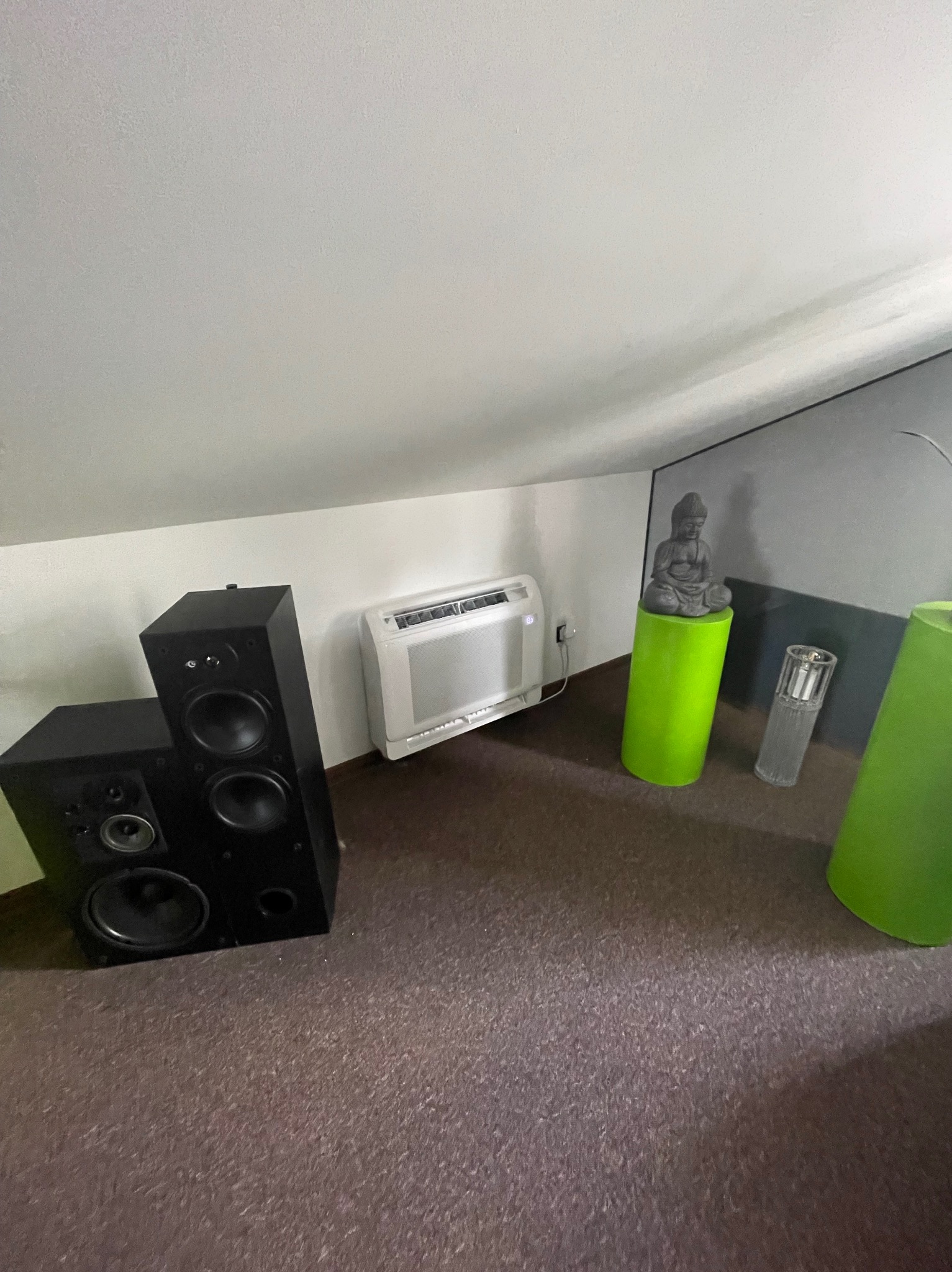 Biały klimatyzator ścienny w pokoju na poddaszu, obok kolumny głośnikowe, zielone dekoracje i statuetka Buddy na zielonym postumencie.