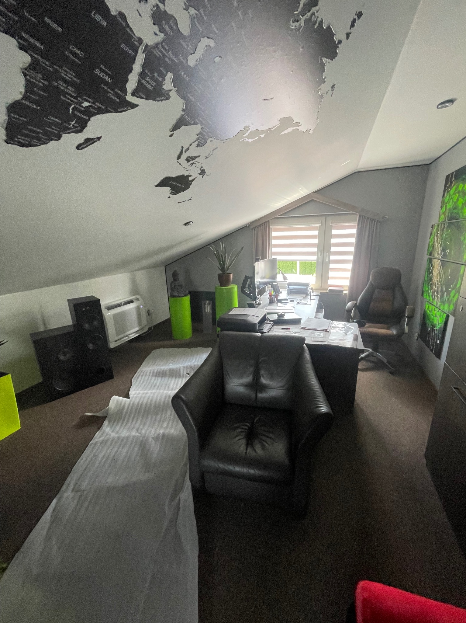 Klimatyzator ścienny w nowoczesnym biurze na poddaszu z mapą świata na suficie, obok głośników i fotela, częściowo przykryty folią ochronną.