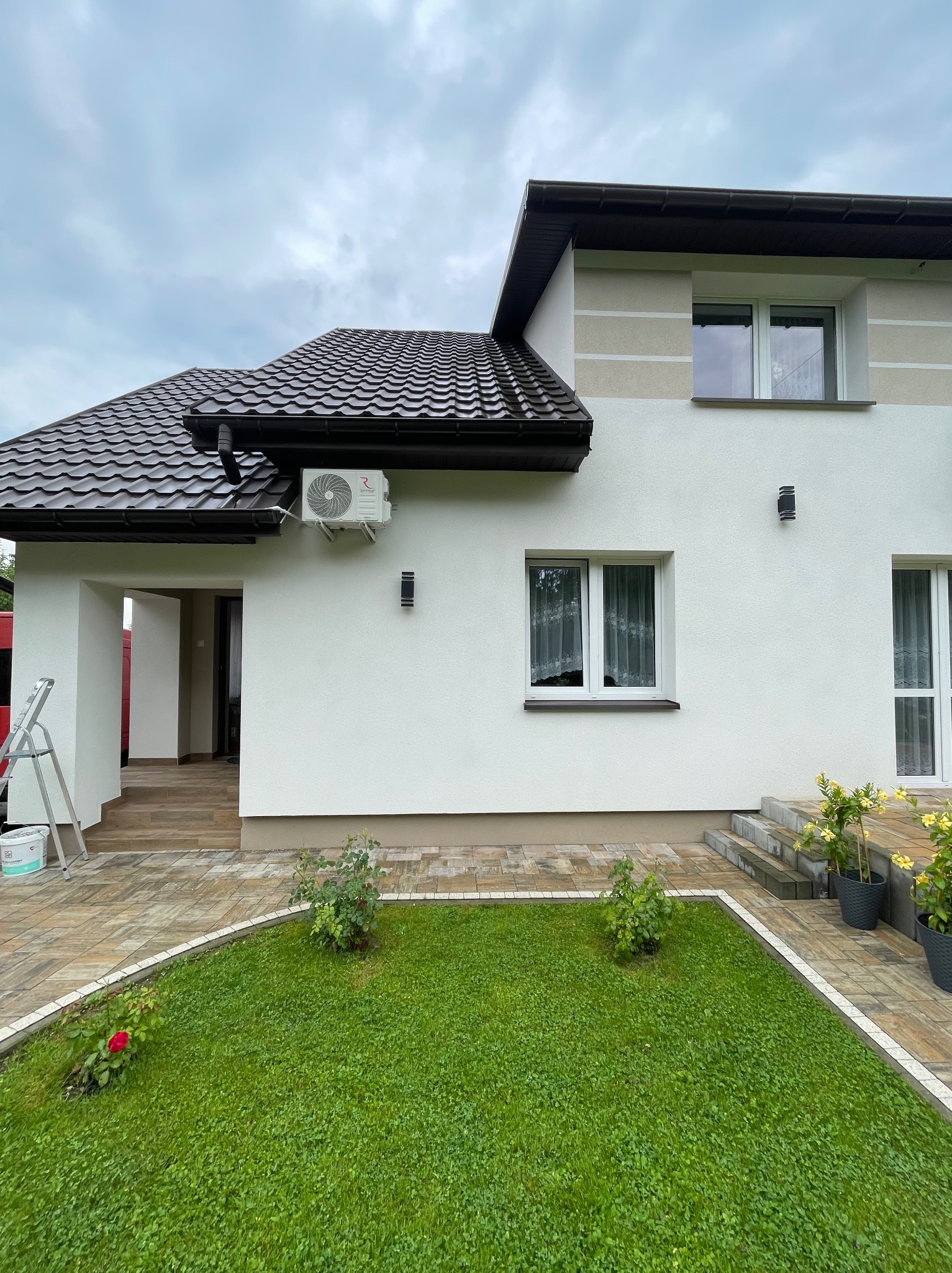 Biały dom z brązowym dachem i zamontowaną klimatyzacją na ścianie. Widoczny ogród z kostką brukową i roślinami. Drabina oparta o ścianę. Pochmurne niebo.