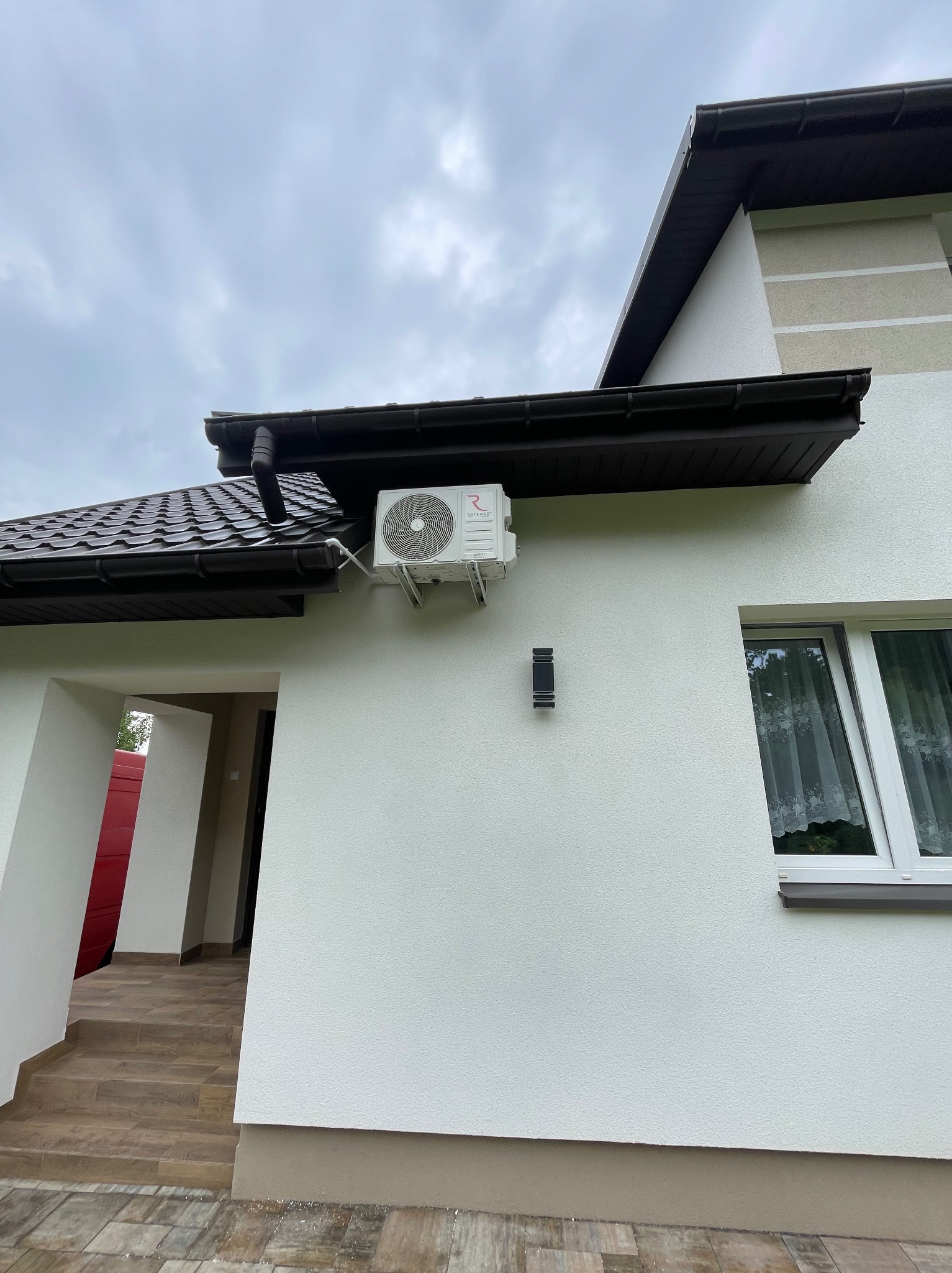 Biała jednostka klimatyzacji zamontowana na ścianie domu z brązowym dachem i oknem z białą ramą, widok z dołu na pochmurne niebo.