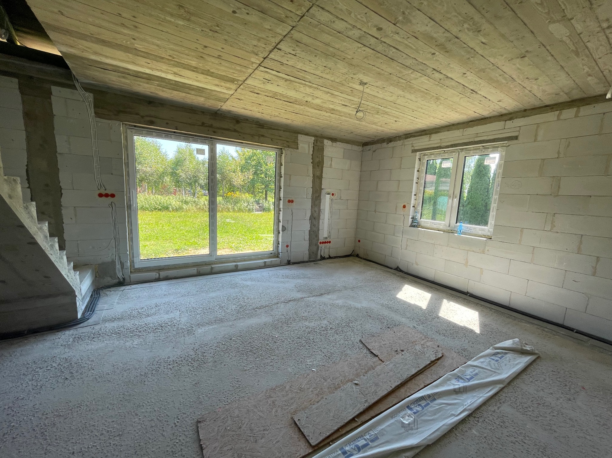 Wnętrze surowego domu w Przysiekach: ściany z betonu komórkowego, okna z widokiem na zieleń, betonowa podłoga i drewniany strop. Widoczne kable elektryczne w ścianach.