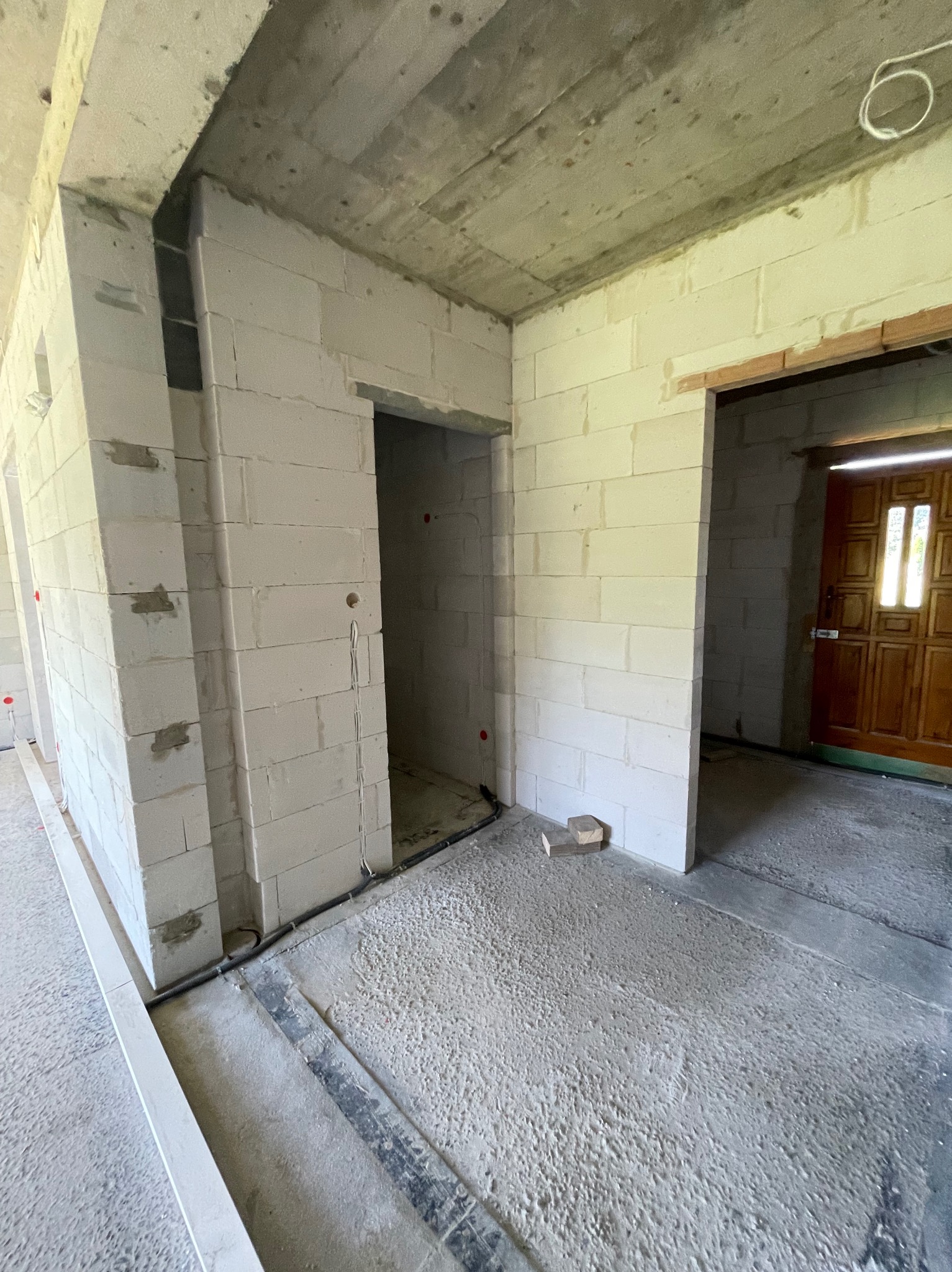 Surowy stan wnętrza: ściany z białych bloczków, widoczne przewody elektryczne i otwory drzwiowe. Podłoga betonowa, sufit surowy, w tle drewniane drzwi.