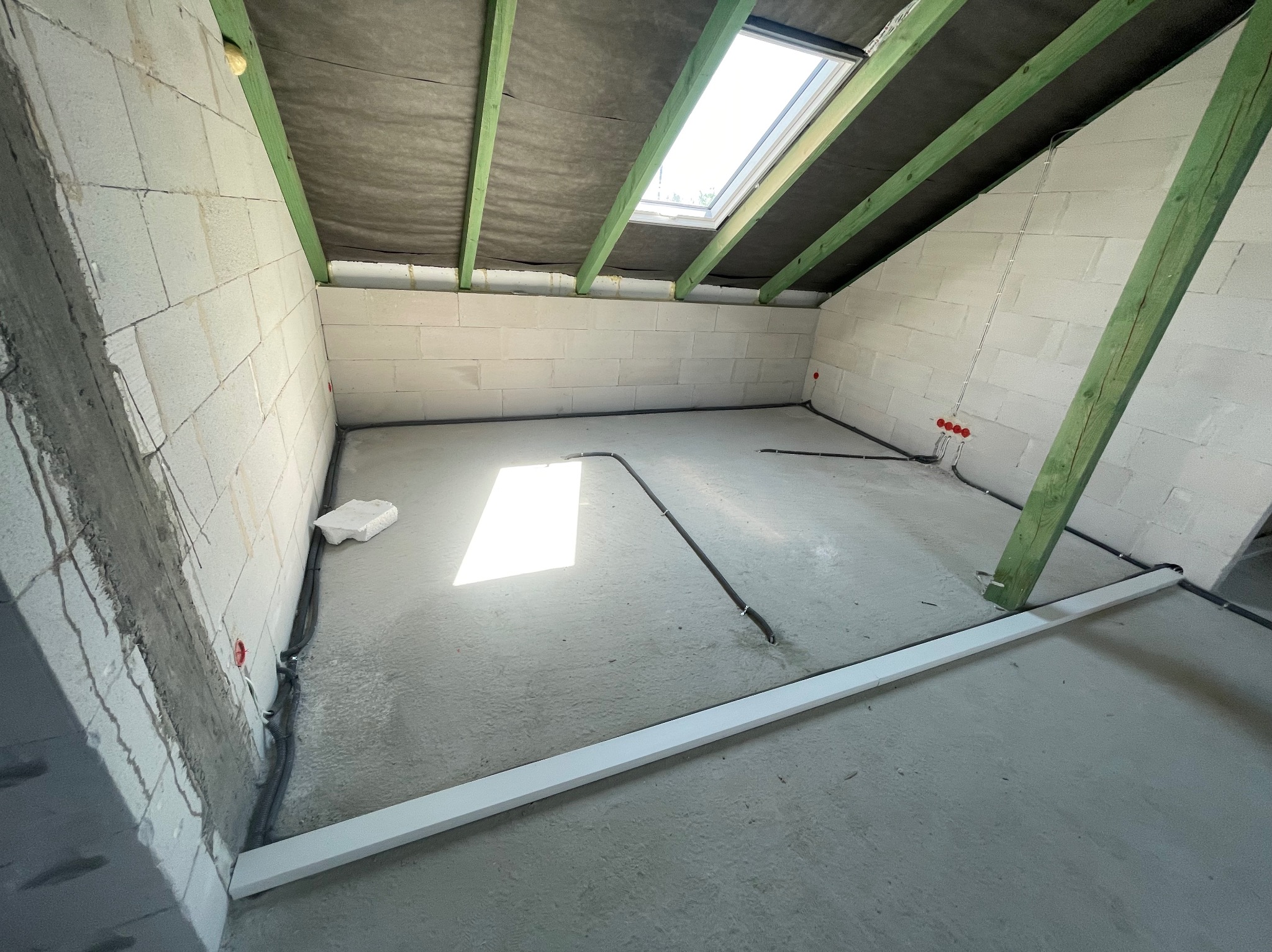 Surowy strych z oknem dachowym, instalacją elektryczną w peszlu i fragmentem listwy przypodłogowej. Widoczne bloczki betonowe i drewniana konstrukcja dachu.