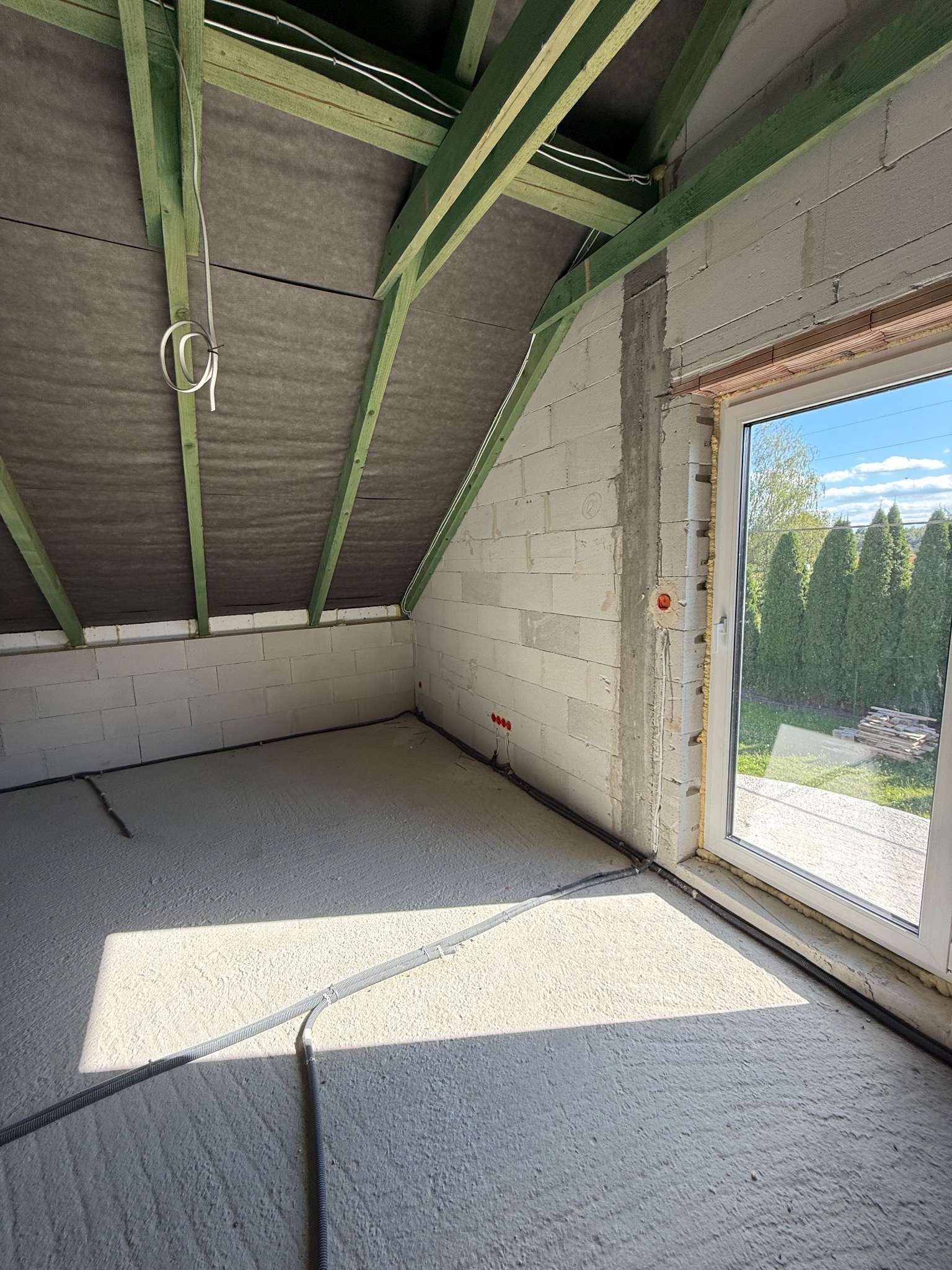 Surowy stan poddasza z widoczną instalacją elektryczną w peszlu przy ścianie i oknach, ściany z betonu komórkowego, drewniana konstrukcja dachu.