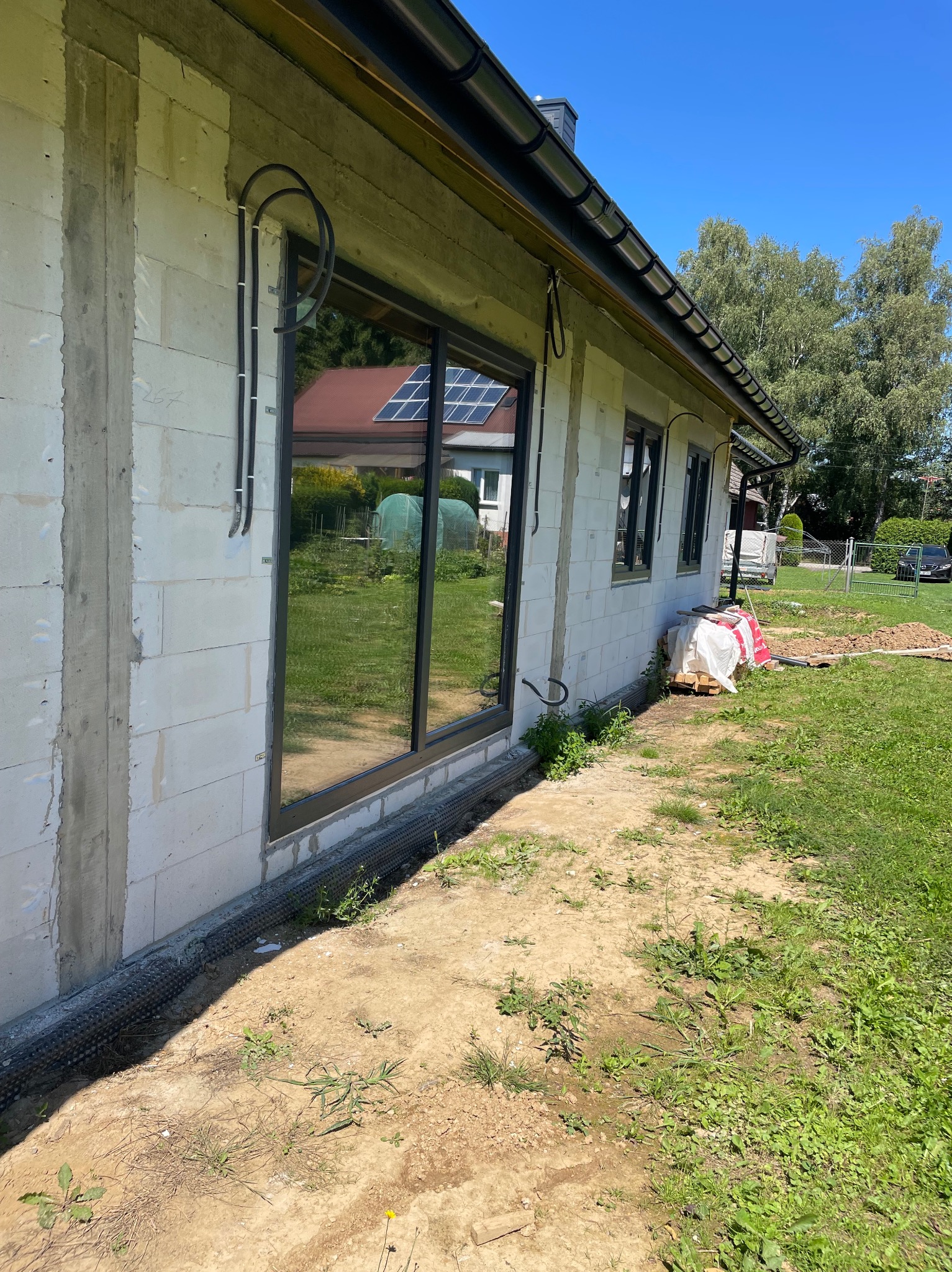 Surowy stan budynku z widocznymi rurami instalacji elektrycznej na elewacji, okno z odbiciem paneli słonecznych w tle, trawa i częściowo zagospodarowany teren.