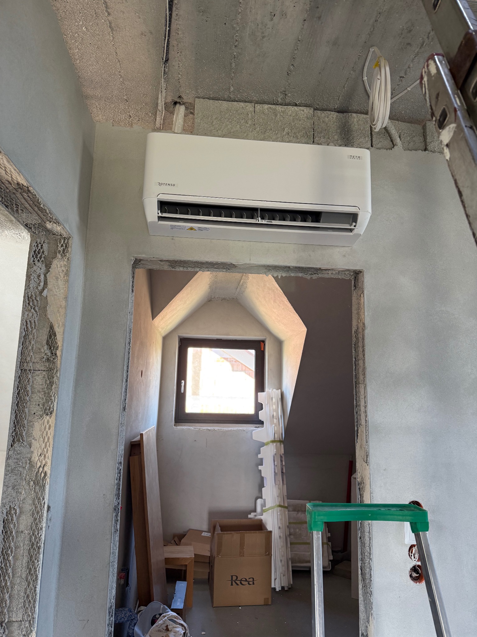 Biały klimatyzator Rotenso Teta zamontowany na szarej ścianie w surowym wnętrzu. Widoczne przewody, drabina i materiały budowlane. Okno w tle.