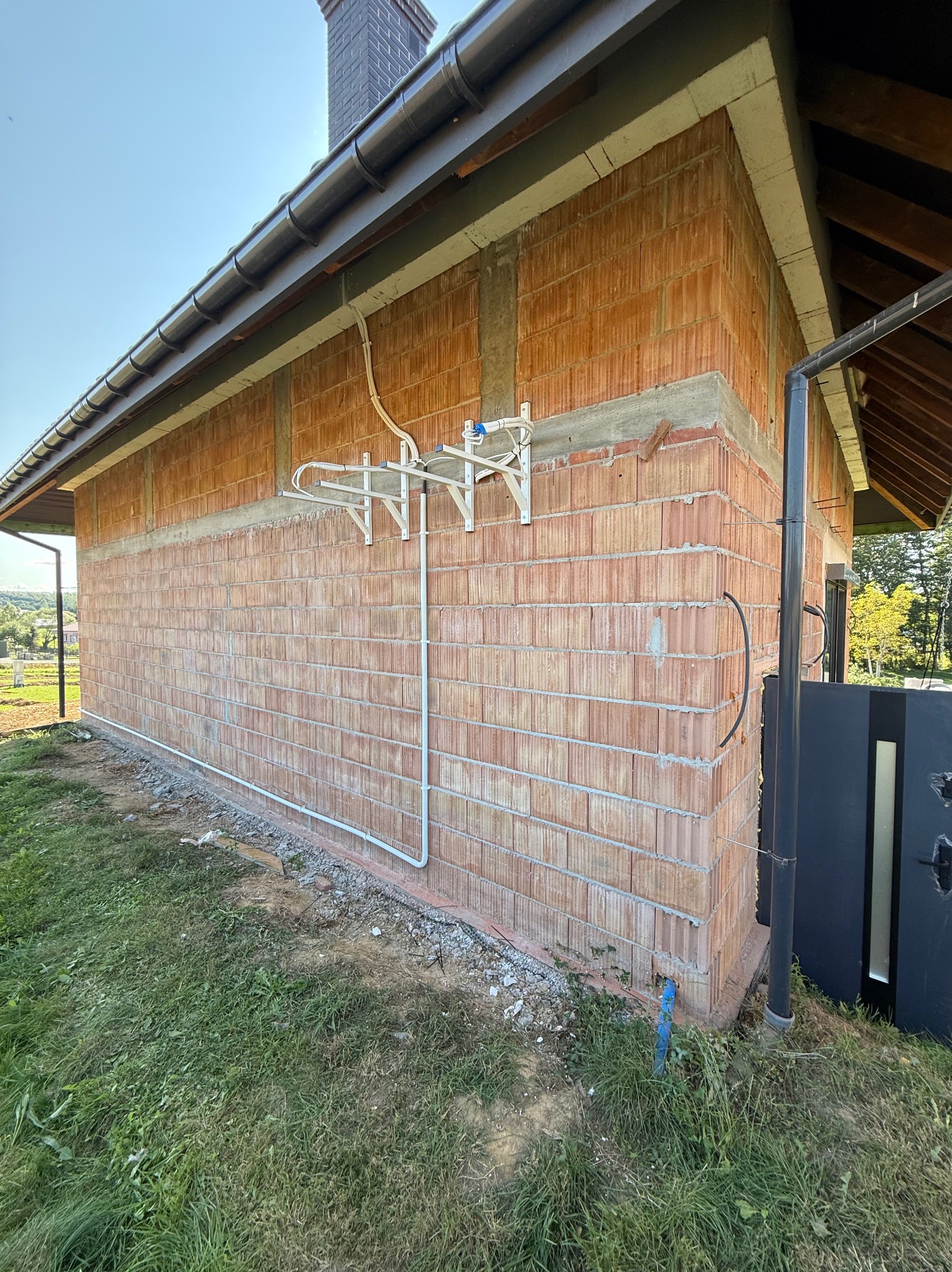 Surowa ściana budynku z cegły z zamontowaną instalacją odgromową i rurami. Widoczne elementy mocujące i przewody. Ujęcie z zewnątrz, trawa w tle.