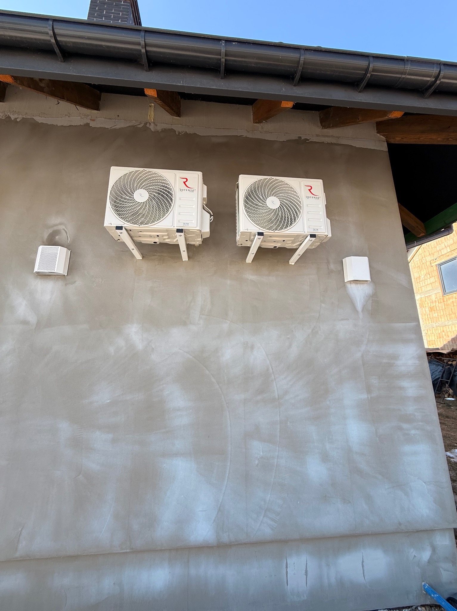 Dwa białe klimatyzatory Rotenso zamontowane na szarej, otynkowanej ścianie budynku. Widoczne elementy instalacji i kratki wentylacyjne. Ujęcie z dołu, błękitne niebo w tle.