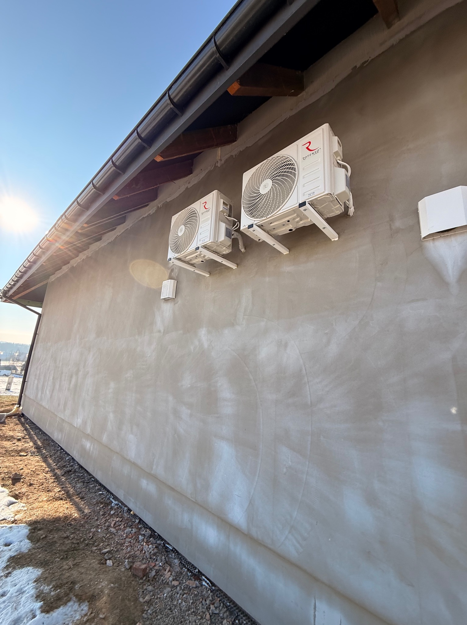 Dwa białe klimatyzatory Rotenso zamontowane na ścianie budynku z surowym tynkiem, widoczne elementy drenażu i orynnowania. Słoneczny dzień.