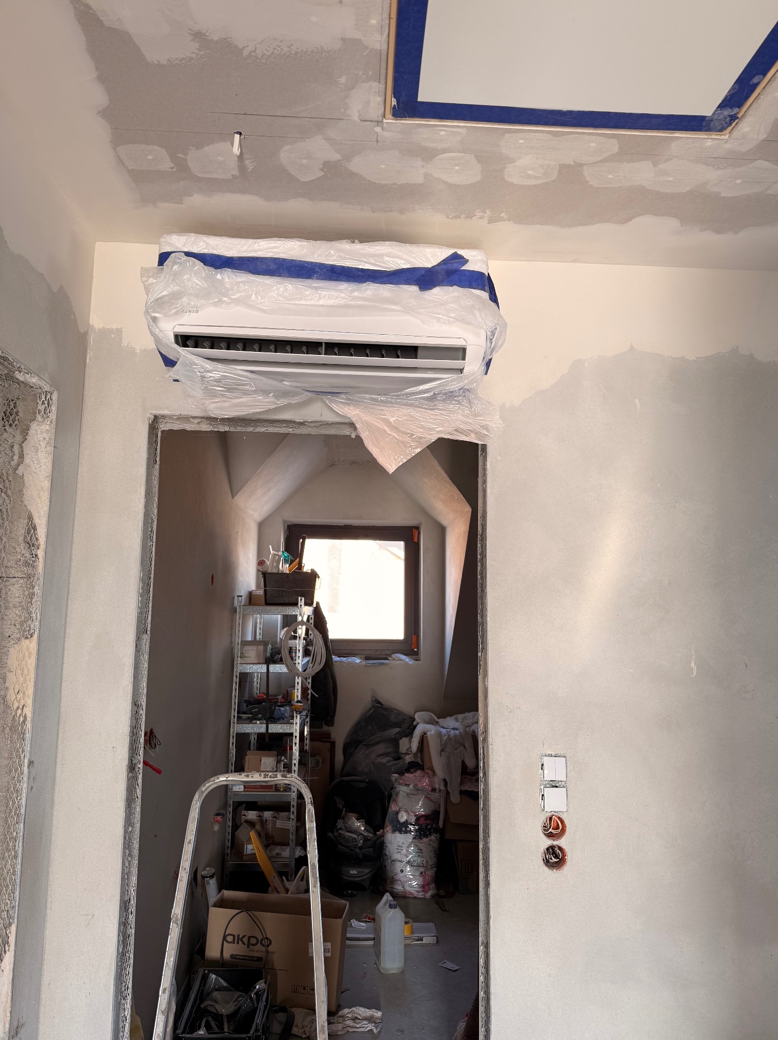 Klimatyzator zabezpieczony folią i taśmą malarską na ścianie w trakcie prac wykończeniowych. Widoczne ślady gładzi i drabina w pomieszczeniu.