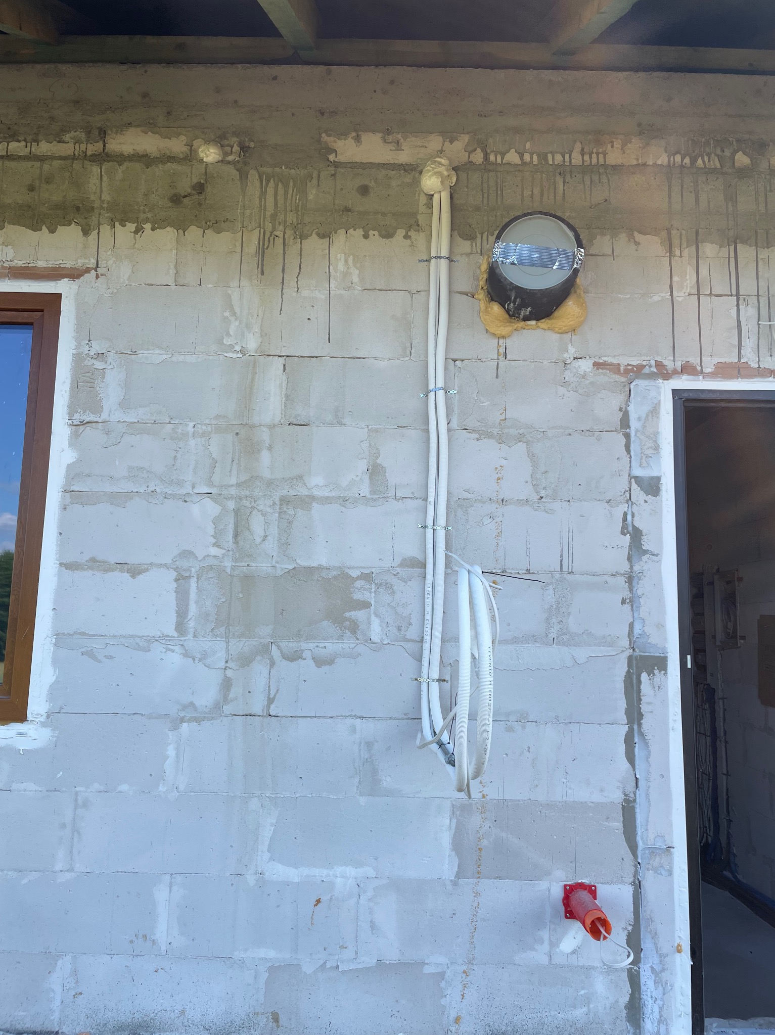 Ściana z białych bloczków z zamontowanymi przewodami elektrycznymi i puszką. Widoczne okno i otwór drzwiowy. Prace instalacyjne w surowym stanie.