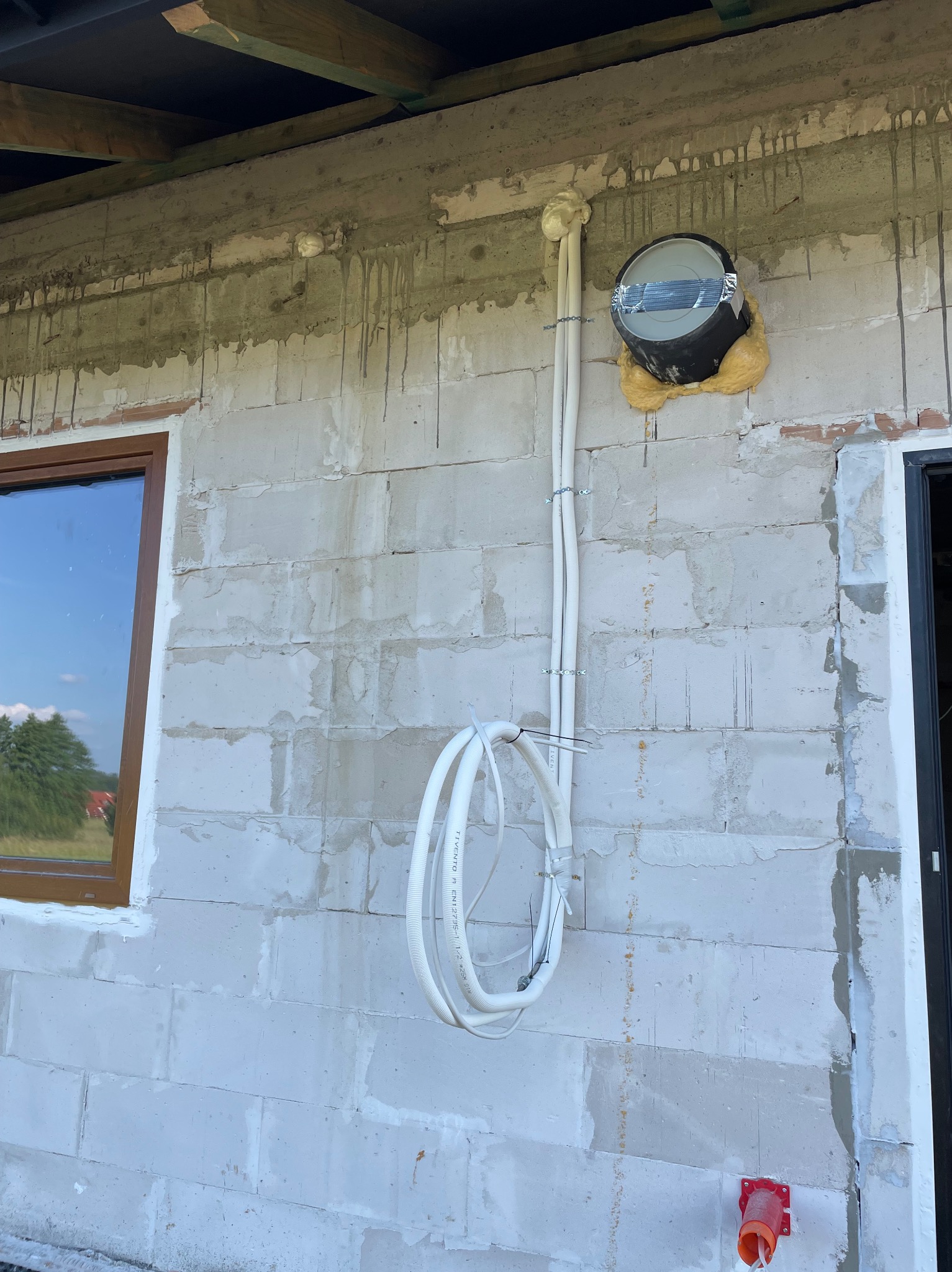 Surowa ściana z białych bloczków z oknem i instalacją elektryczną w peszlu, zakończoną puszką. Widoczne ślady po zaprawie i pianka montażowa.