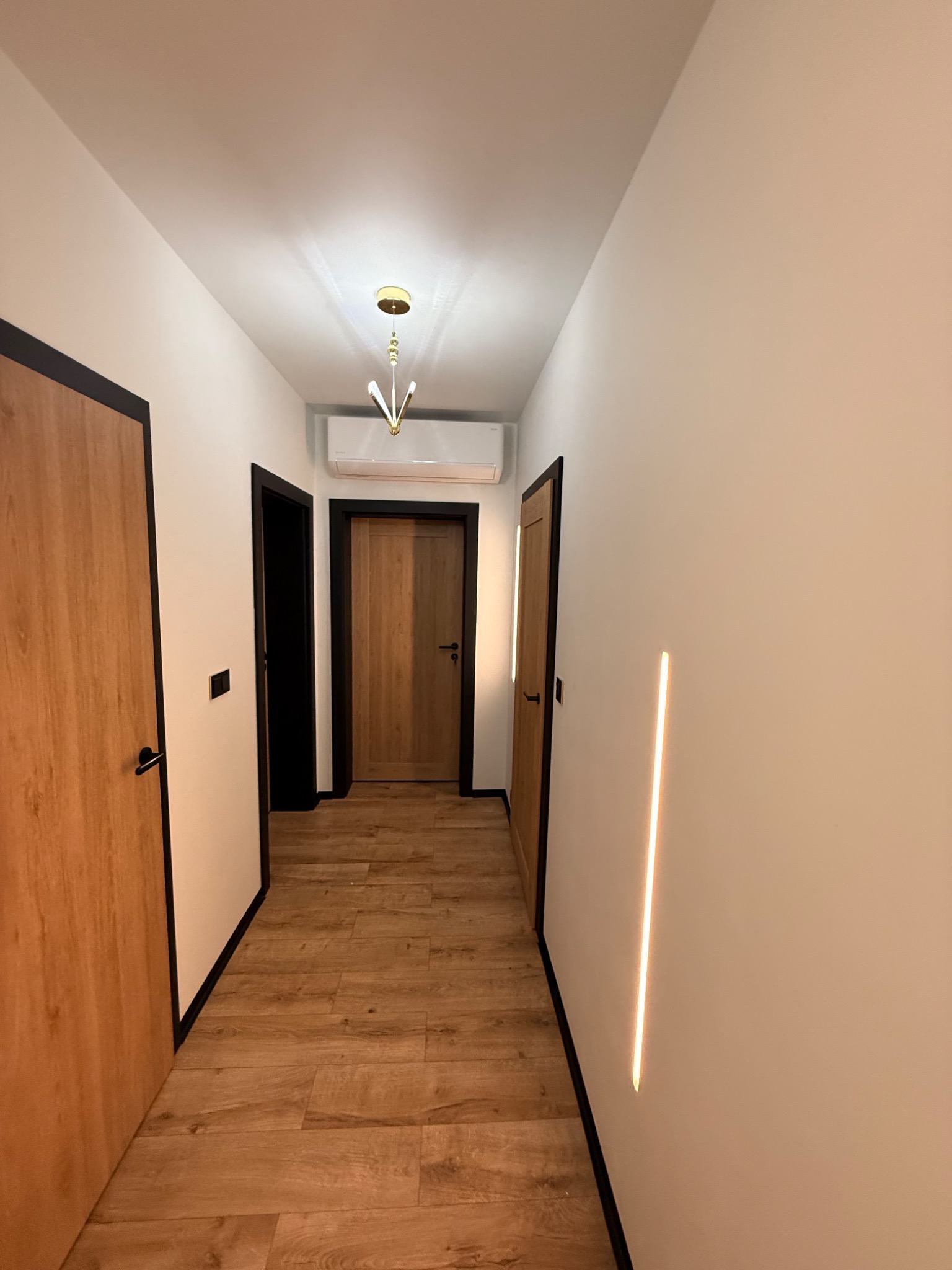 Korytarz z drewnianymi drzwiami z czarnymi ramami, podświetlana listwa LED w ścianie, klimatyzator nad drzwiami i złoty żyrandol.