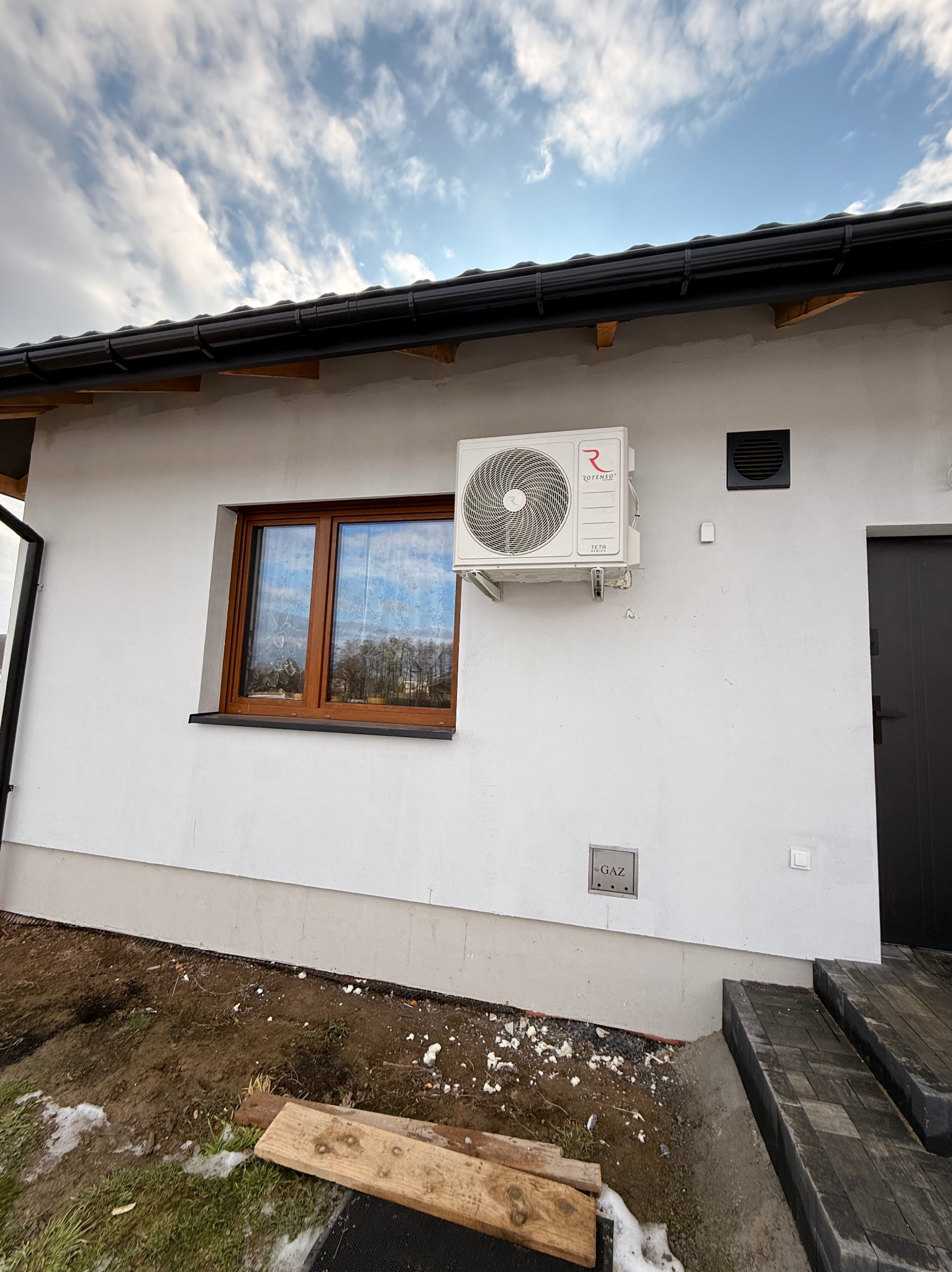 Biała jednostka zewnętrzna klimatyzacji Rotenso Teta zamontowana na elewacji domu z brązowymi oknami i czarną rynną. Widoczna skrzynka gazowa.