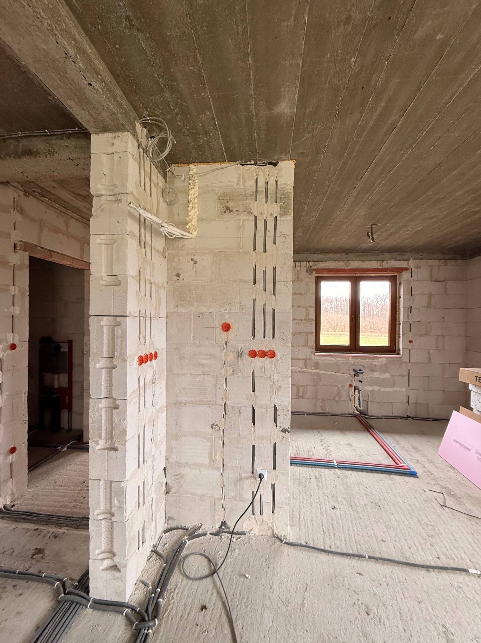 Surowy stan wnętrza z widoczną instalacją elektryczną w ścianach z betonu komórkowego, okablowaniem pod sufitem i rurami instalacji wodnej na podłodze.