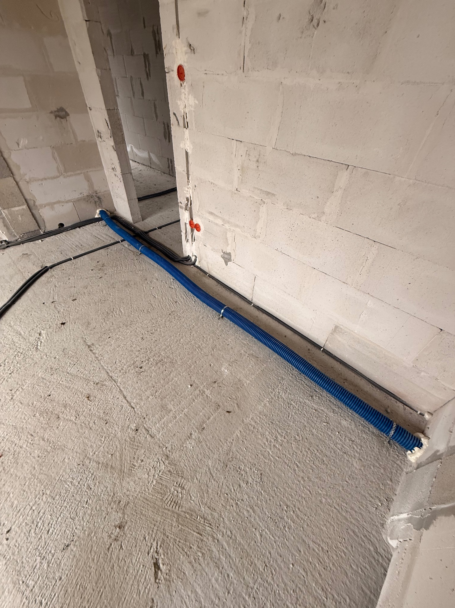 Niebieska rura karbowana przymocowana do ściany w surowym wnętrzu. Widoczne kable i puszki elektryczne. Ściany z betonu komórkowego.