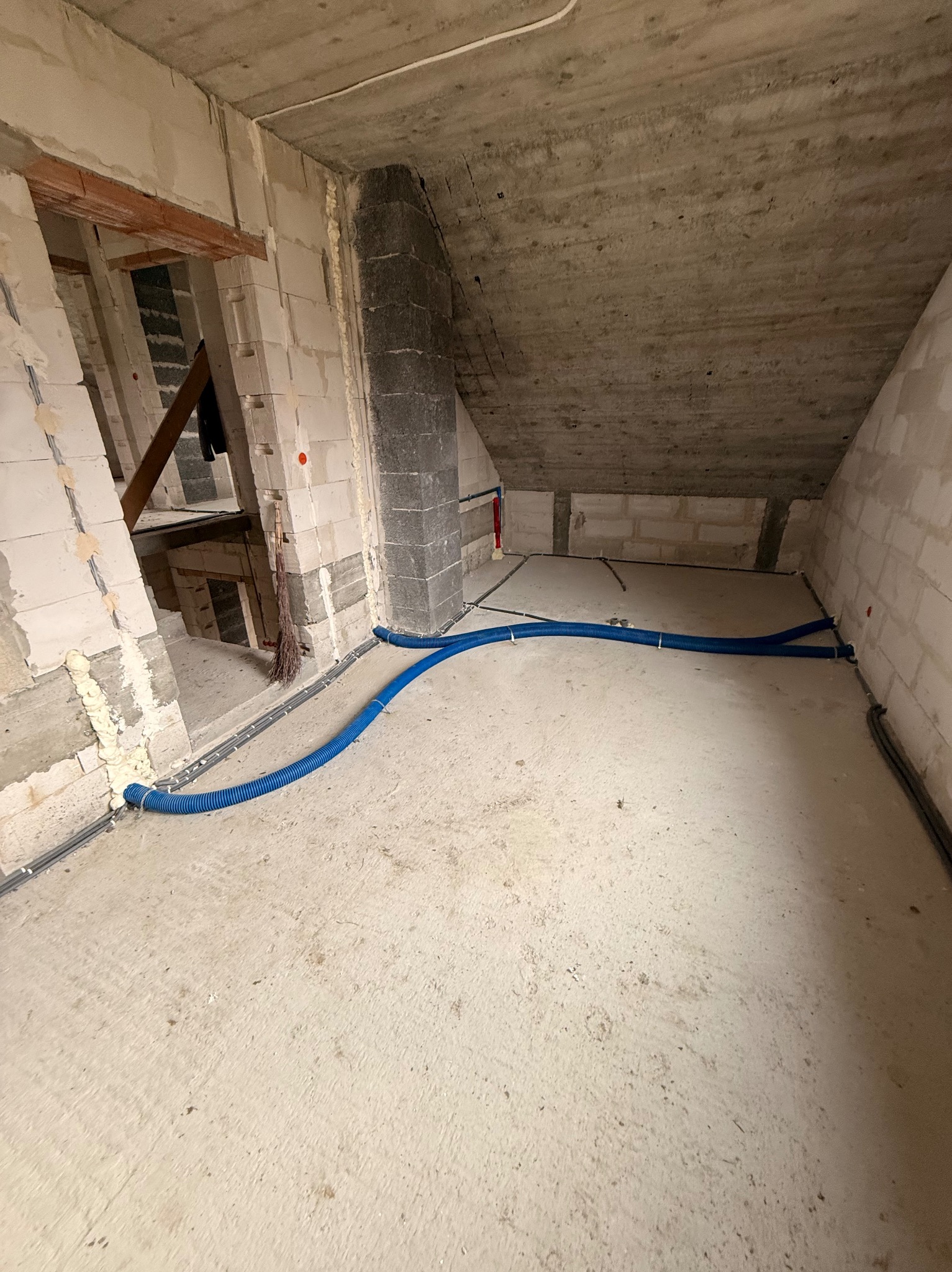 Surowy stan poddasza z widoczną instalacją w niebieskiej karbowanej rurze, prowadzoną po podłodze wzdłuż ścian. Widoczne przewody elektryczne i fragmenty ścian z pustaków.