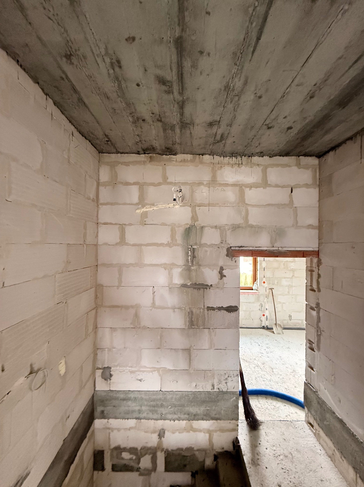 Surowy stan wnętrza z białych bloczków, widoczne przewody instalacji elektrycznej i oznaczenia 'KLIMA' na ścianie, sufit betonowy, w tle widoczne narzędzia budowlane.