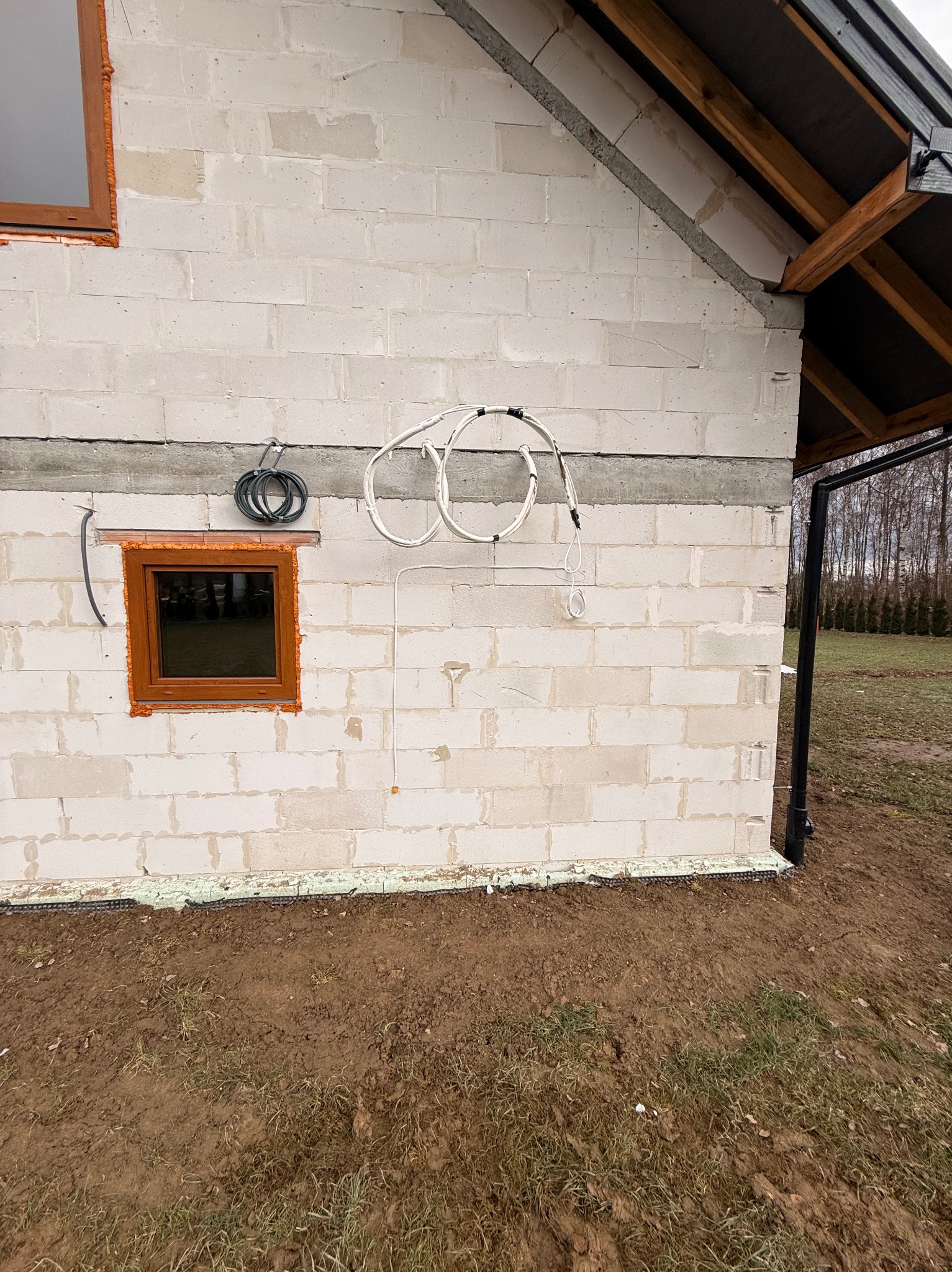 Ściana budynku w trakcie budowy z widocznymi przewodami elektrycznymi i oknem z brązową ramą. Widoczne elementy instalacji na zewnątrz budynku.