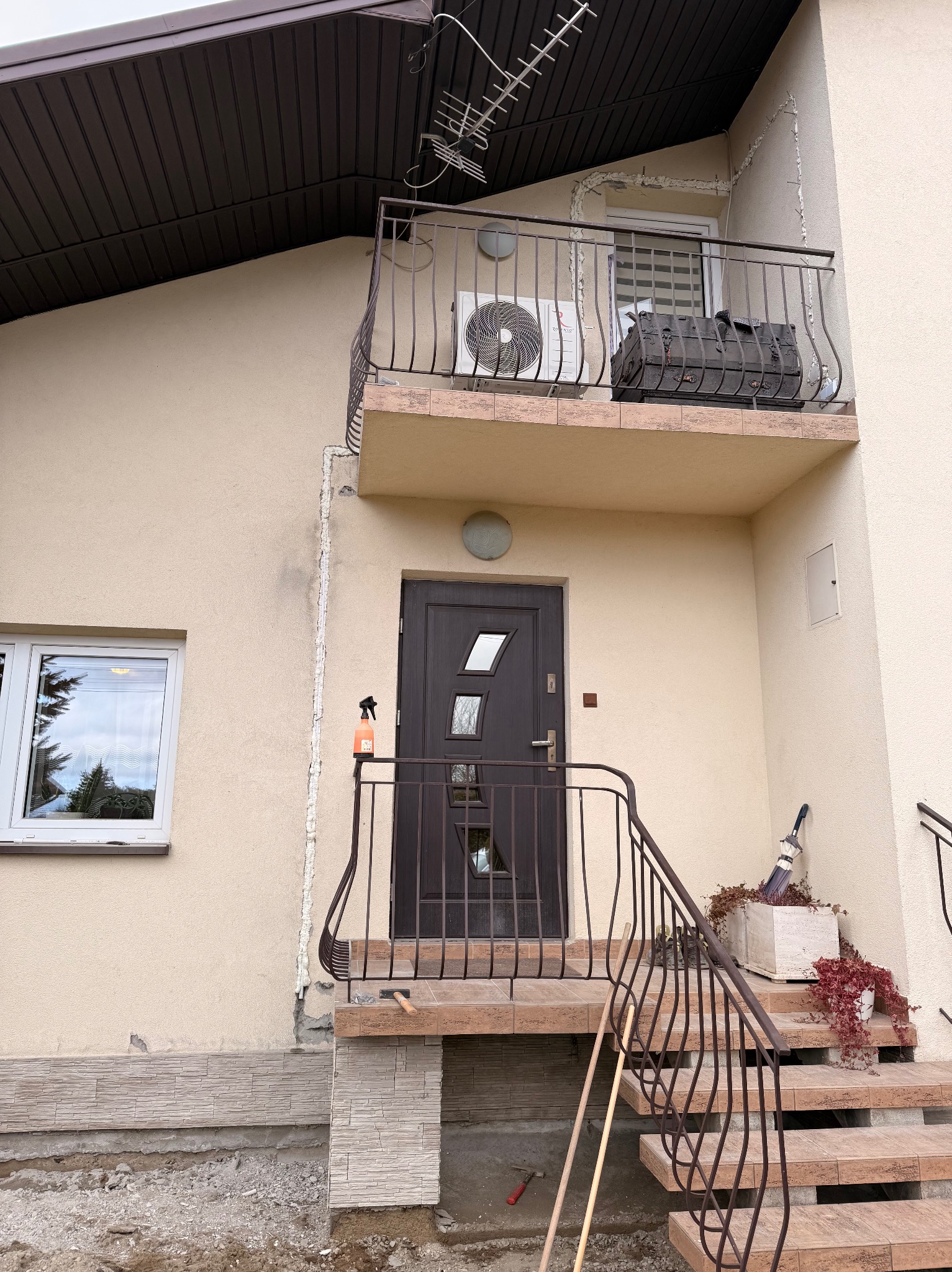 Dom z balkonem, na którym zamontowano klimatyzator. Widoczne schody wejściowe z metalową barierką i narzędzia. Elewacja budynku w jasnym odcieniu.