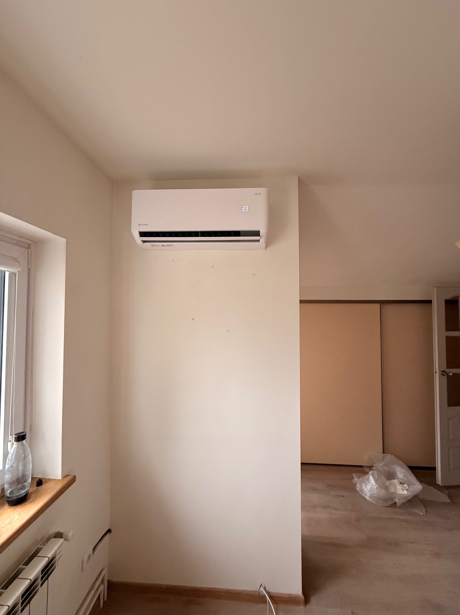 Biały klimatyzator ścienny marki Totenso zamontowany w jasnym pomieszczeniu z oknem i grzejnikiem. Widoczny fragment podłogi i drzwi do innego pokoju.