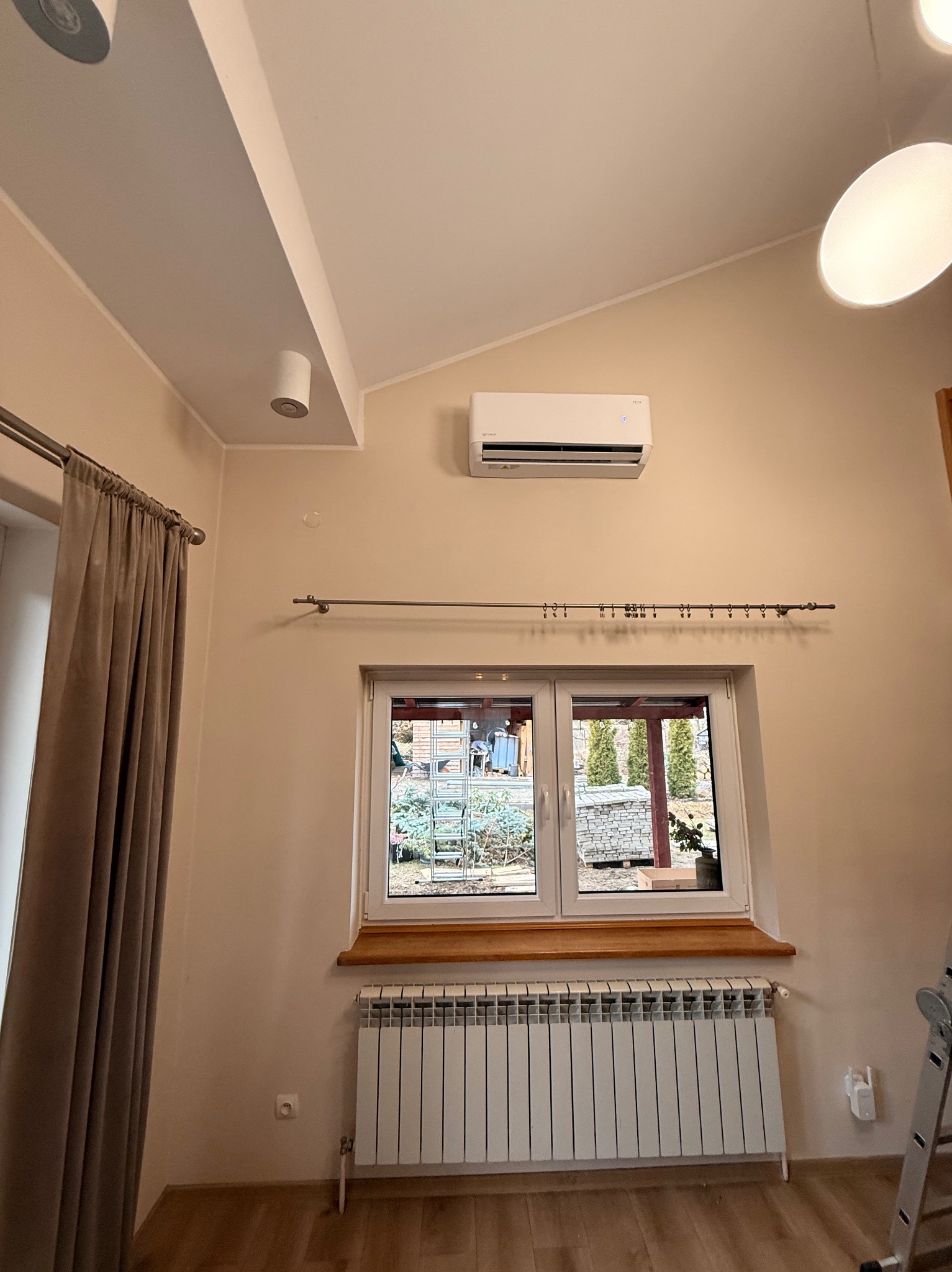 Biały klimatyzator na ścianie w jasnym pokoju z oknem i grzejnikiem. Widoczne elementy instalacji elektrycznej i oświetlenia.