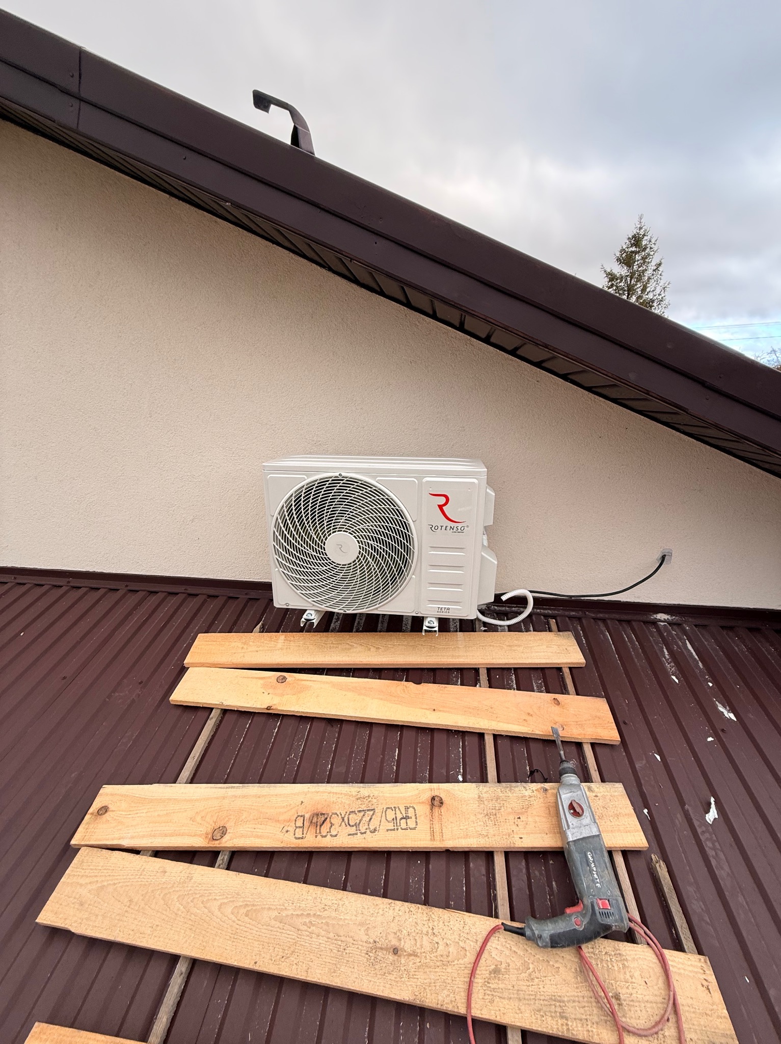 Biała jednostka zewnętrzna klimatyzacji Rotenso zamontowana na brązowym dachu, z widocznymi deskami i wiertarką, sugerująca trwający montaż.