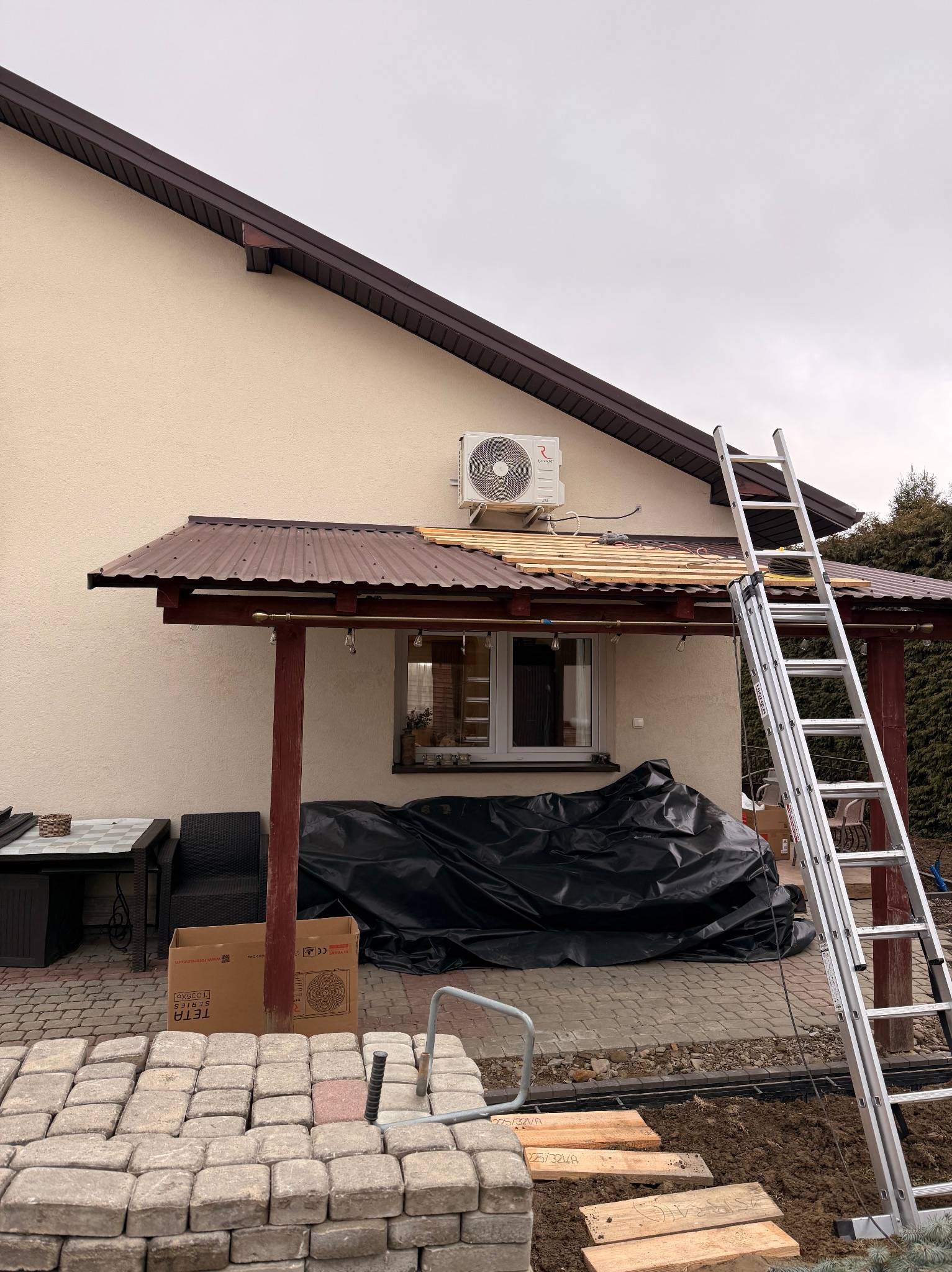 Montaż klimatyzacji zewnętrznej na elewacji domu, widoczna drabina oparta o dach ganku, kostka brukowa i karton po klimatyzatorze na pierwszym planie.