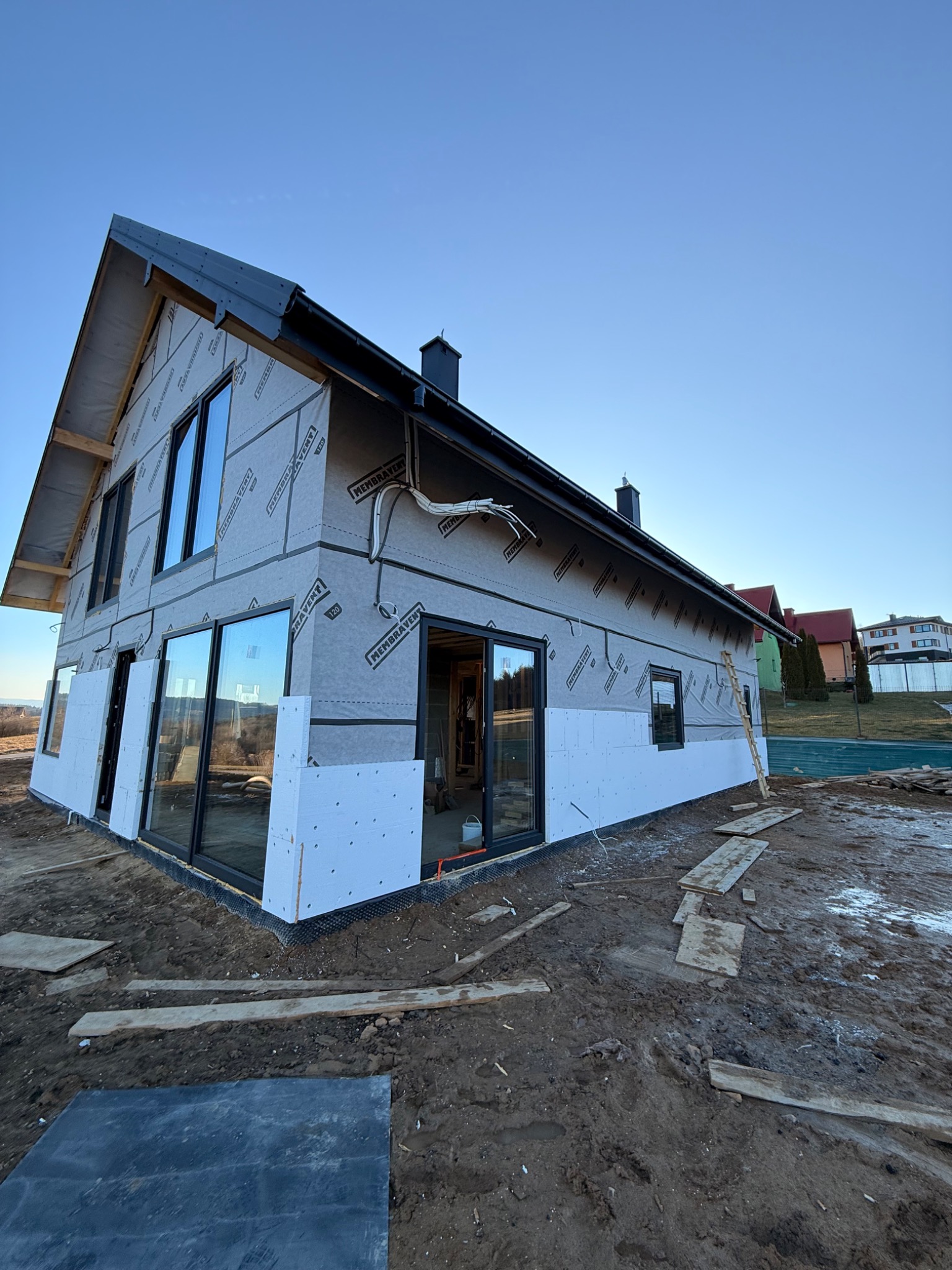 Dom jednorodzinny w trakcie ocieplania, widoczne płyty izolacyjne i folia wiatroizolacyjna. Stolarka okienna w kolorze czarnym. Teren wokół domu rozkopany.