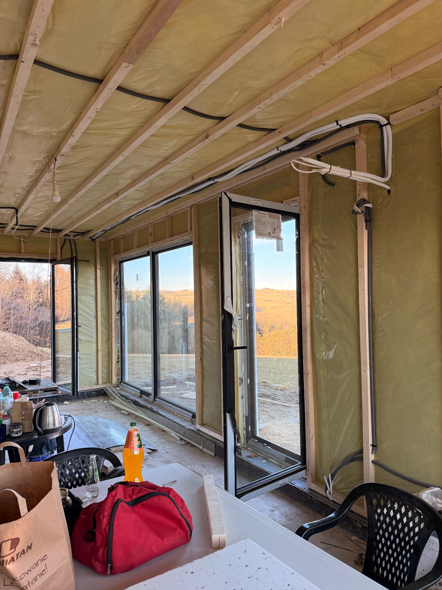 Wnętrze w trakcie prac wykończeniowych: widoczne ocieplone ściany, drewniana konstrukcja, okna z widokiem na krajobraz i instalacje elektryczne w peszlach.