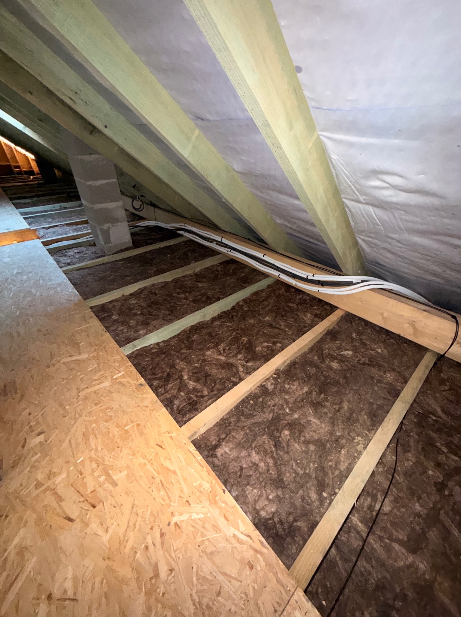 Ocieplenie poddasza wełną mineralną, widoczne drewniane krokwie i instalacja elektryczna w peszlach. Częściowo ułożona płyta OSB.