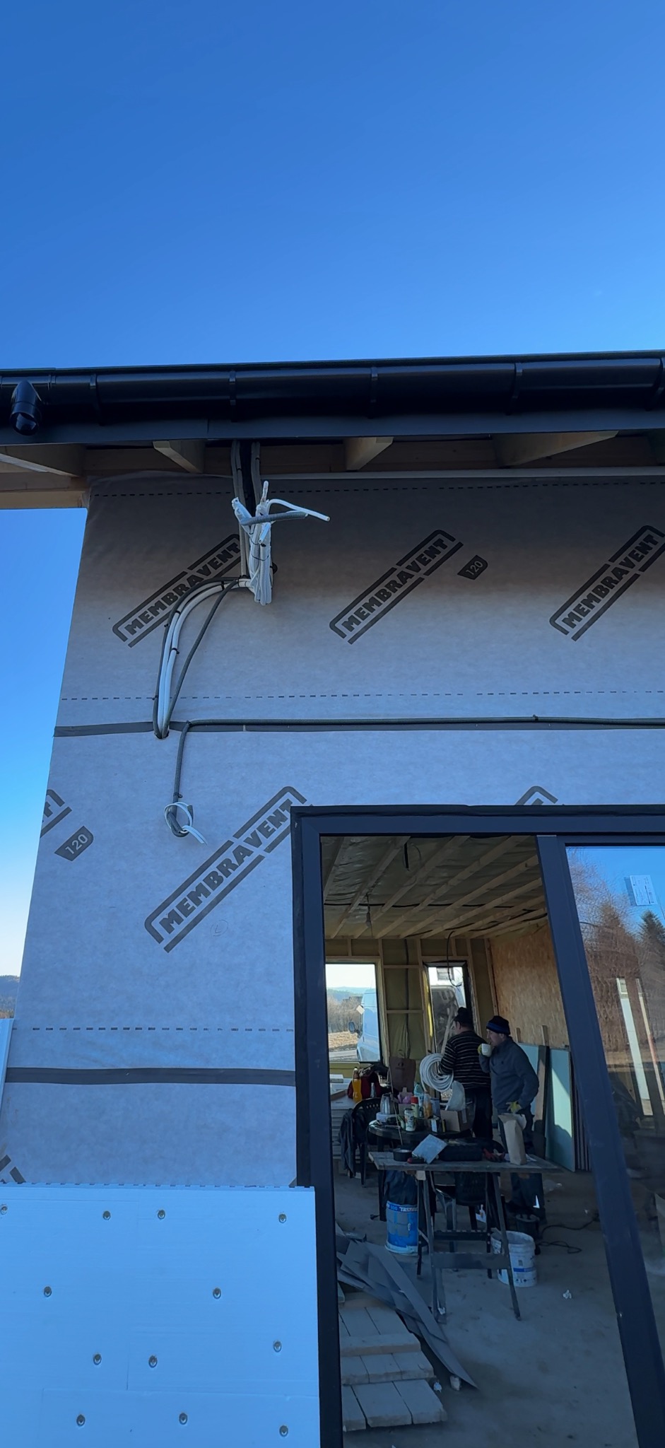 Montaż instalacji elektrycznej na zewnątrz budynku w trakcie budowy. Widoczne kable i przewody przymocowane do ściany z membraną. W tle ekipa budowlana.