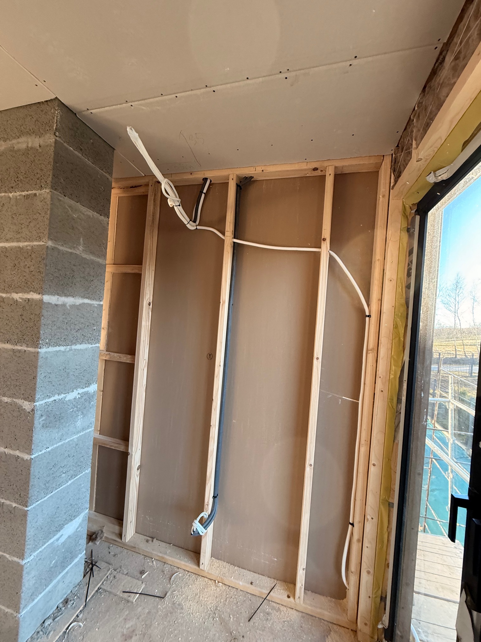 Instalacja elektryczna w trakcie montażu w Jasło. Widoczne przewody w ścianie szkieletowej, płyty gipsowe na suficie i fragment okna z widokiem na zewnątrz.