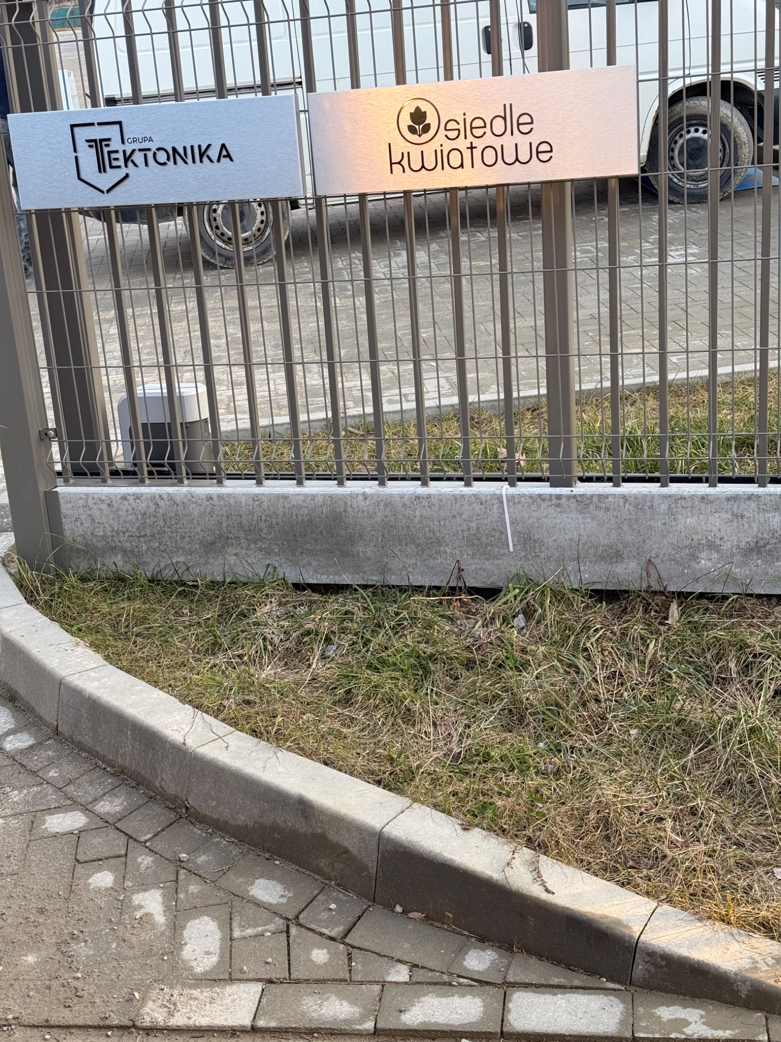 Tabliczki z nazwami firm 'Tektonika' i 'Siedle Kwiatowe' umieszczone na metalowym ogrodzeniu, z fragmentem zaparkowanego białego busa w tle.