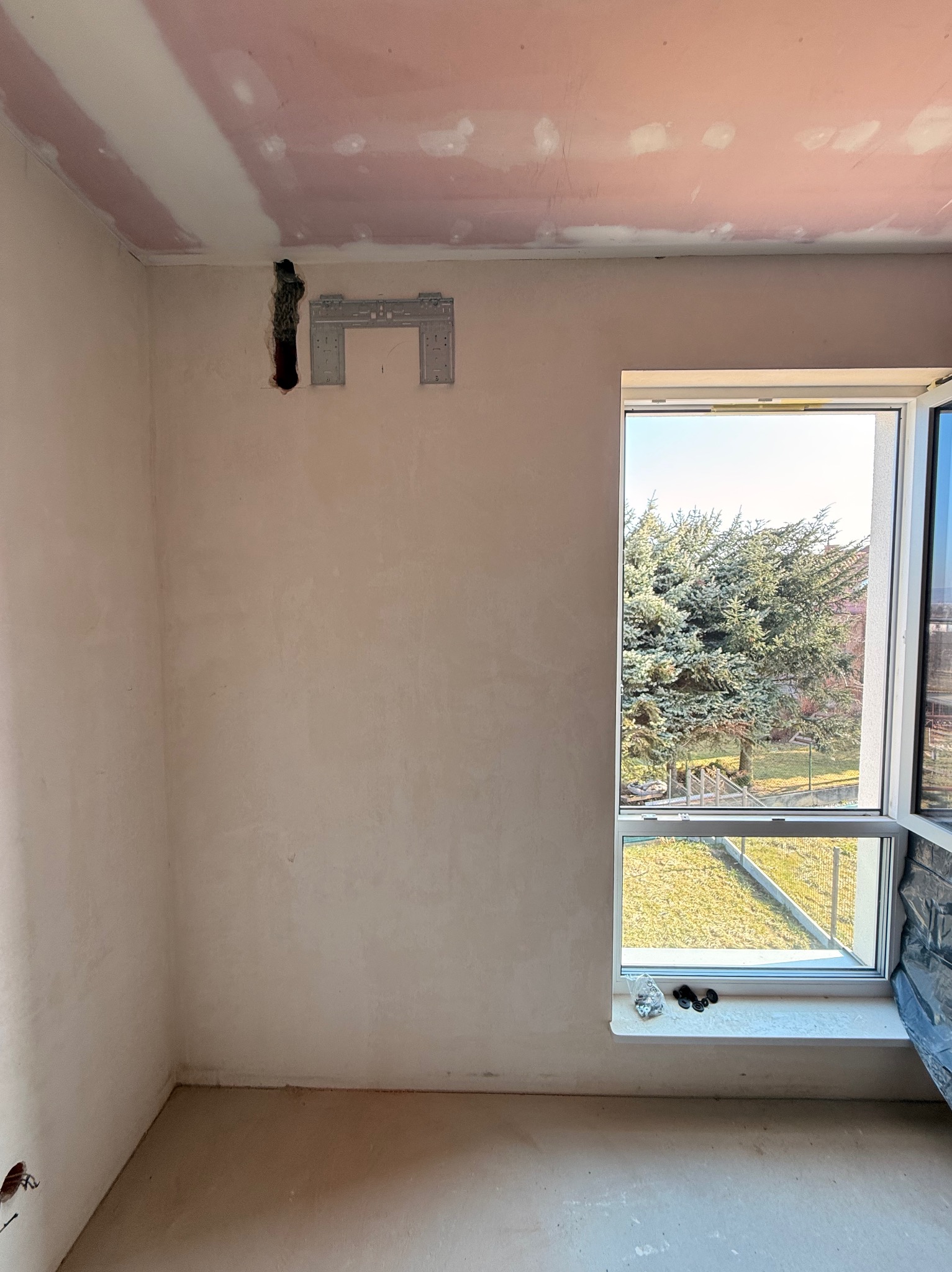 Wnętrze w trakcie remontu: przygotowane miejsce na klimatyzator z zamontowanym uchwytem, widoczne otwory w ścianie, okno z widokiem na zieleń.