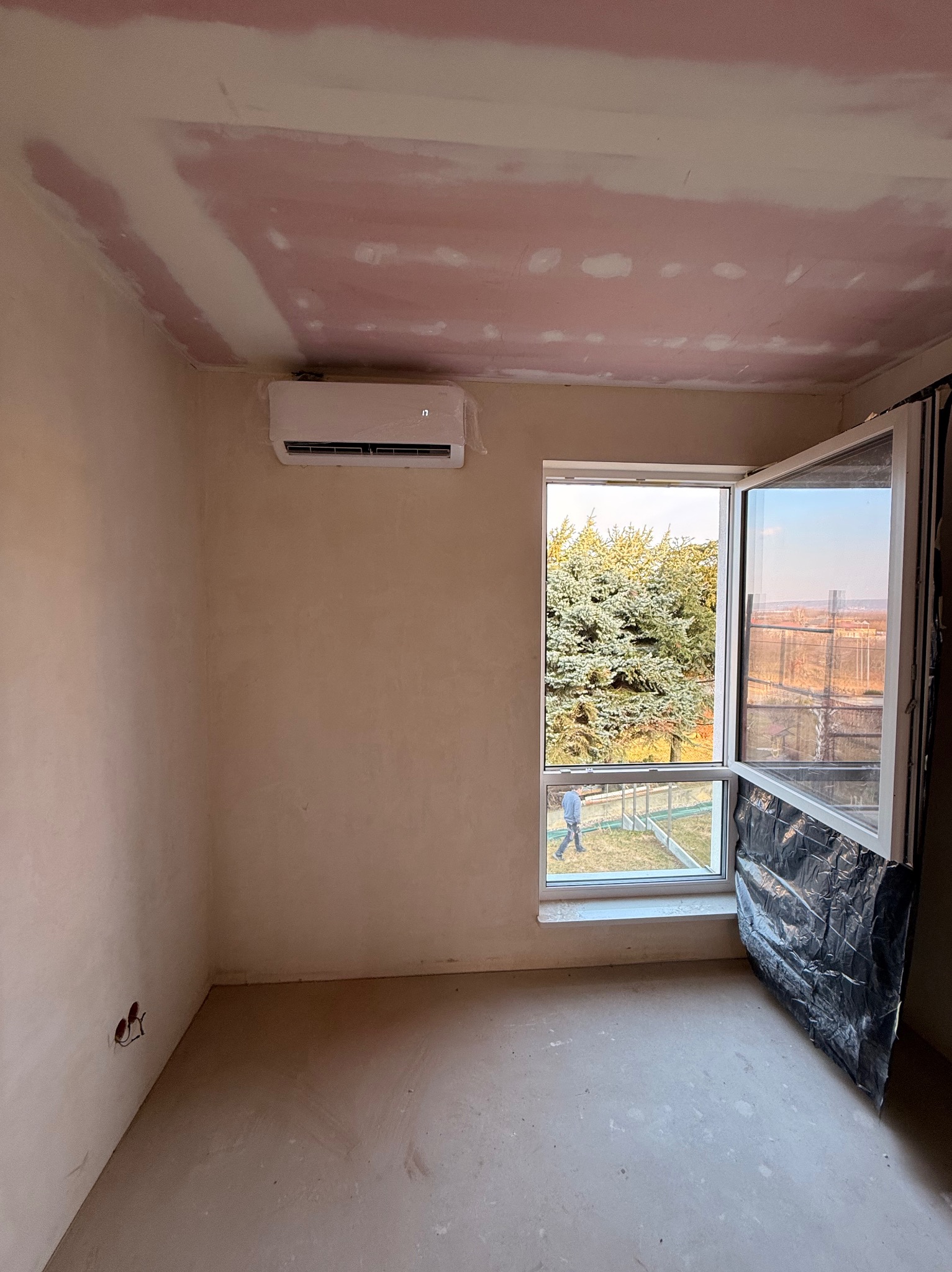 Klimatyzator ścienny zamontowany w surowym wnętrzu w Jaśle. Widoczne przygotowania do wykończenia ścian i sufitu. Otwarta okno z widokiem na zewnątrz.