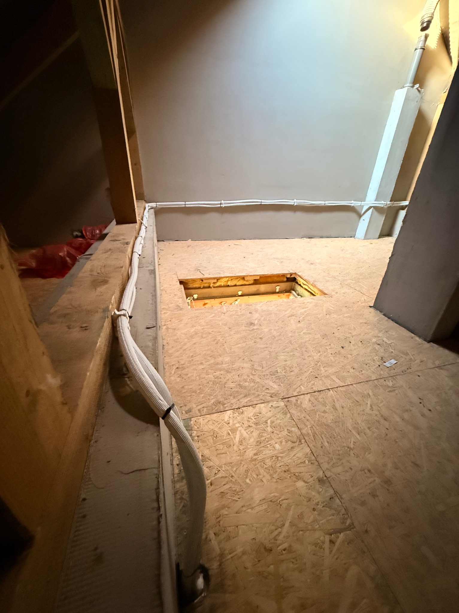 Instalacja kabli w osłonie na poddaszu, widoczne elementy konstrukcyjne i otwór w podłodze. Białe kable prowadzone wzdłuż ściany, jasne oświetlenie.
